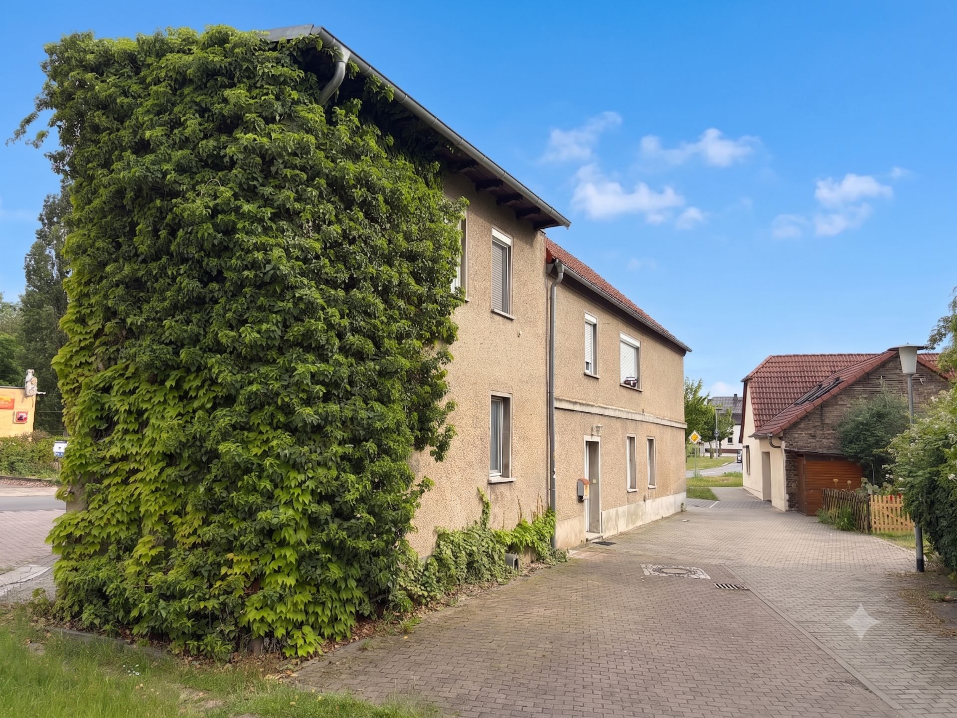 Zweistöckiges Wohnhaus mit zugewachsener Fassade und grauem Gehweg davor.