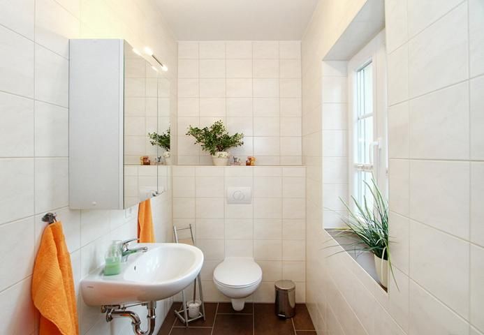 Helles Badezimmer mit Waschbecken, WC, Spiegel, orangenen Handtüchern und Pflanzen am Fenster.