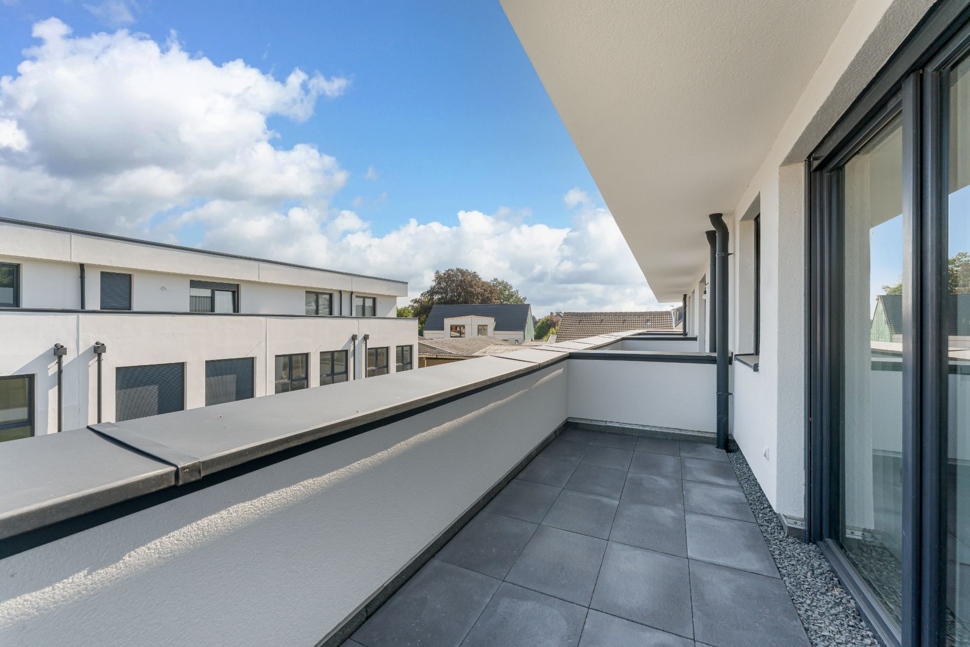 Großer Balkon mit Fliesen, Glasgeländer und Blick auf benachbarte Wohngebäude.