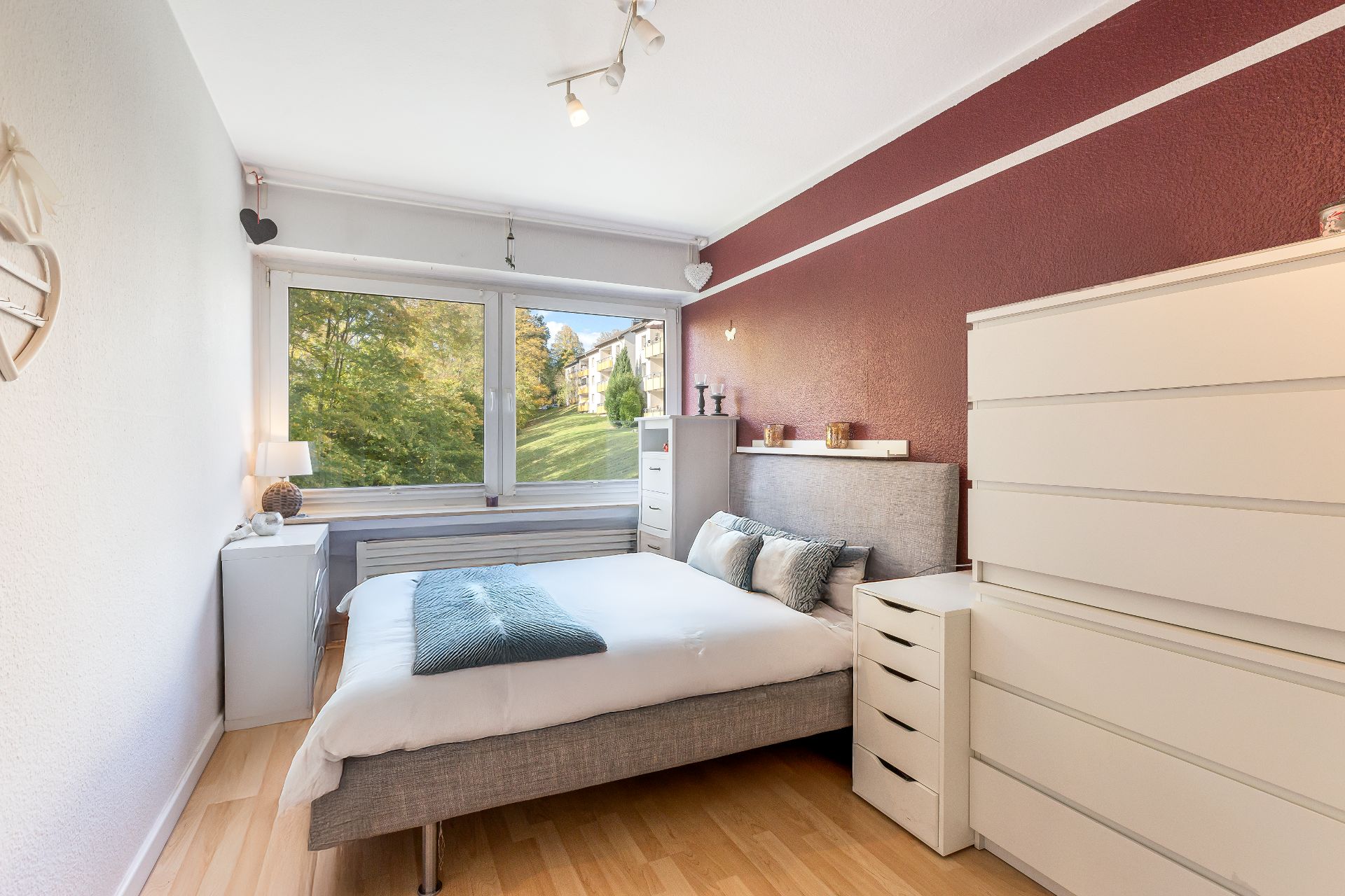 Helles Schlafzimmer mit großem Fenster, Doppelbett und weißen Kommoden auf Laminatboden.