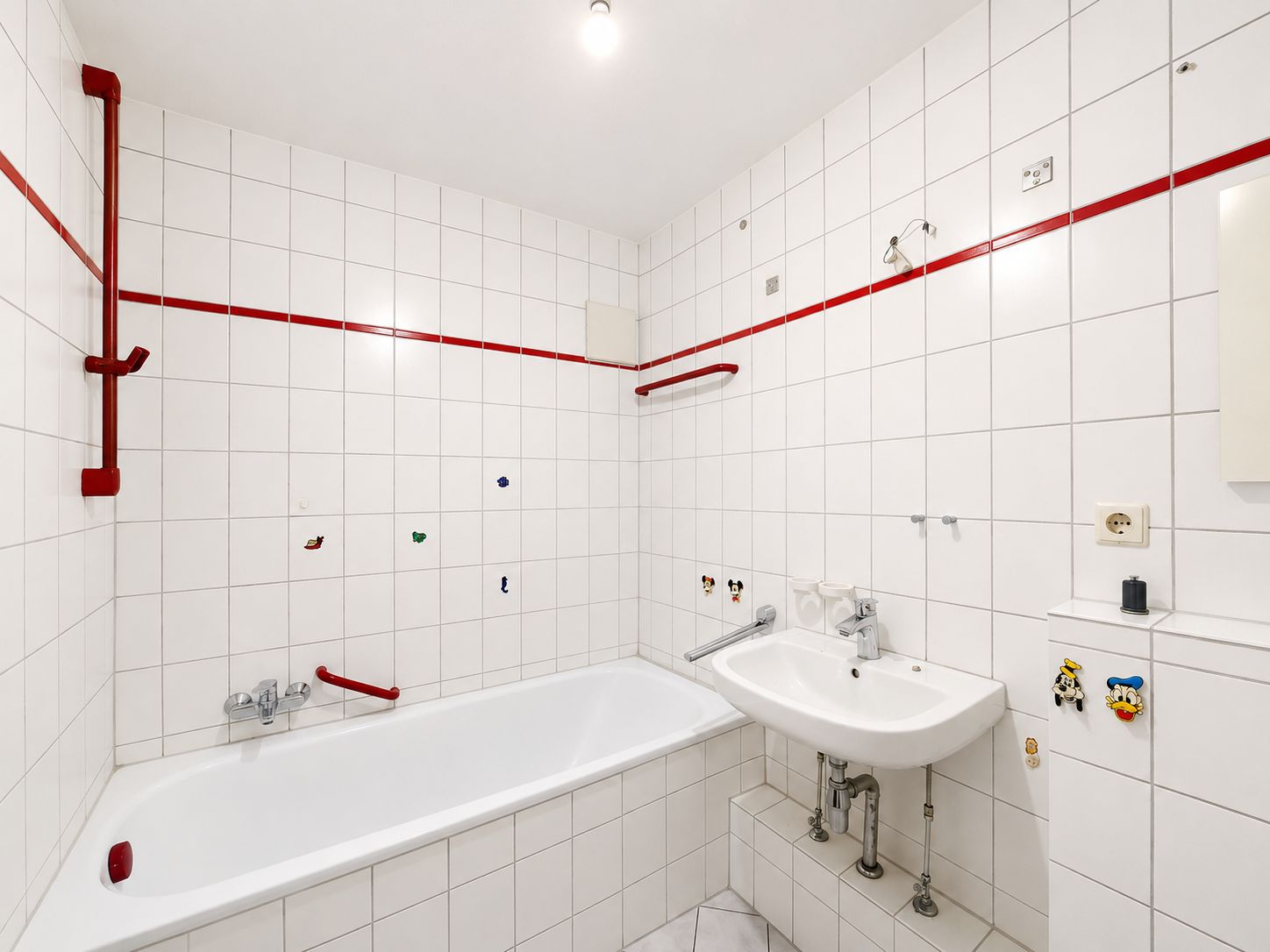 Weiß gefliestes Badezimmer mit Badewanne, Waschbecken und roten Haltegriffen.