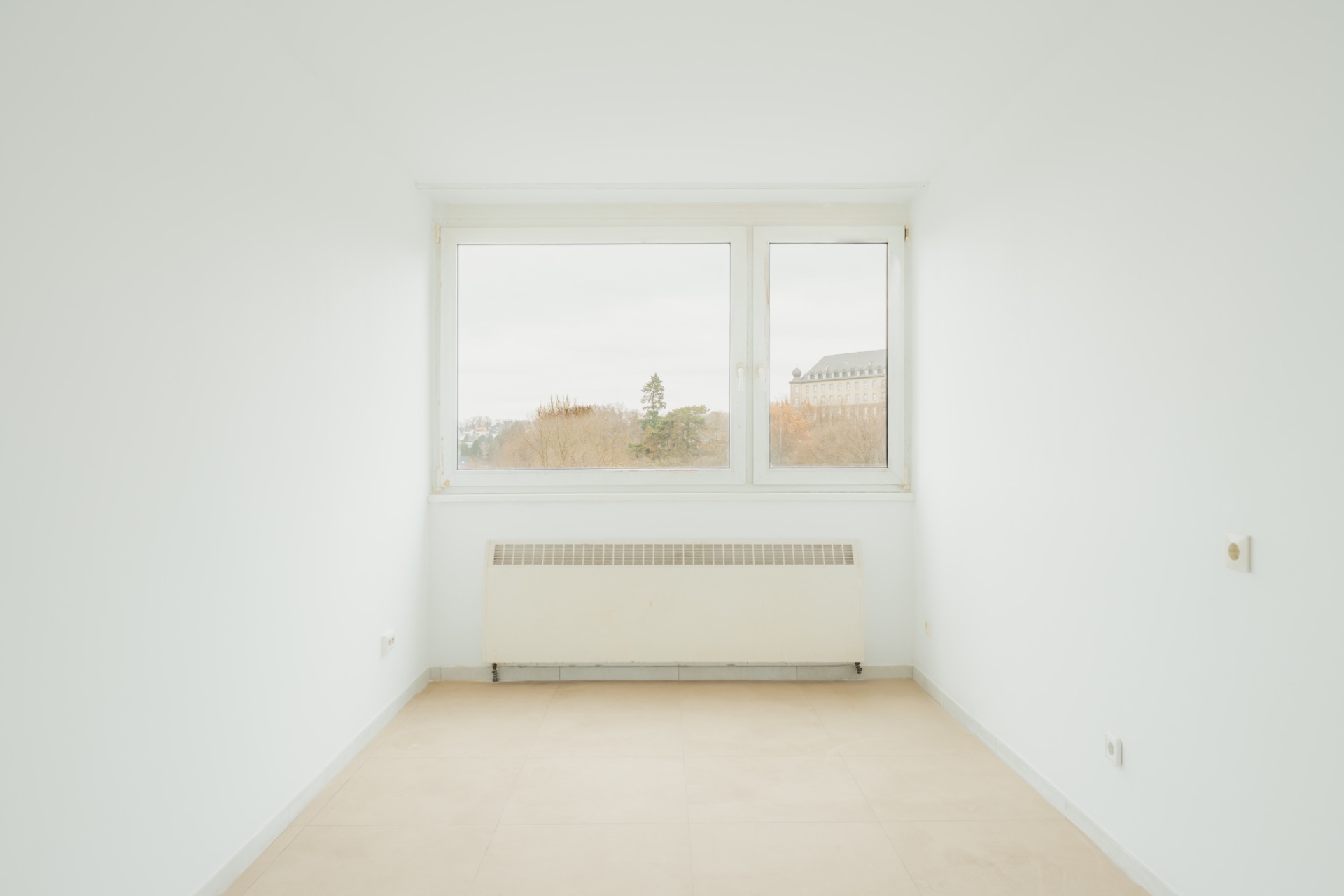 Helles, leeres Zimmer mit großem Fenster und Heizkörper an der Wand.