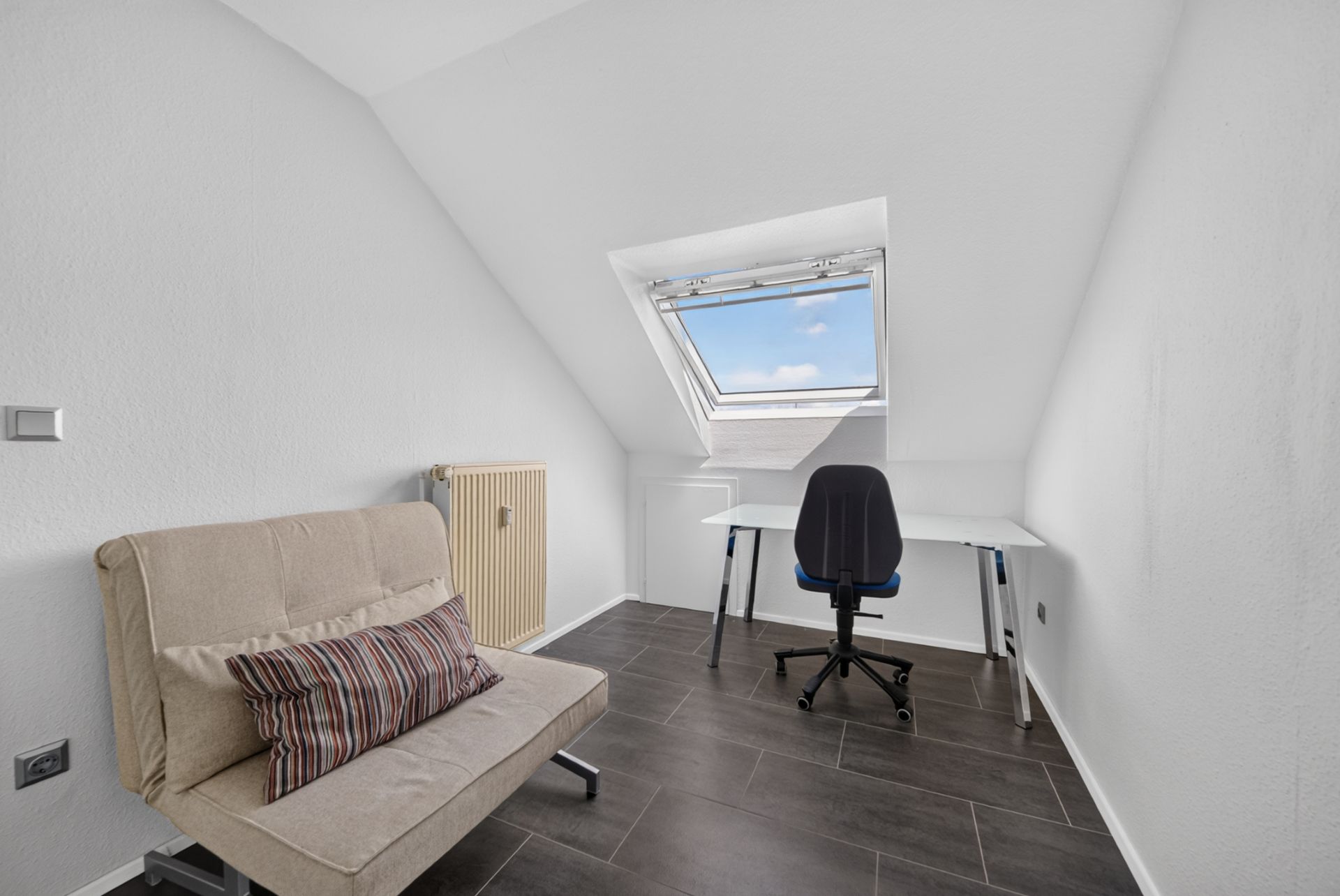 Helles Zimmer mit Schlafsofa, Schreibtisch und Stuhl unter einem Dachfenster.
