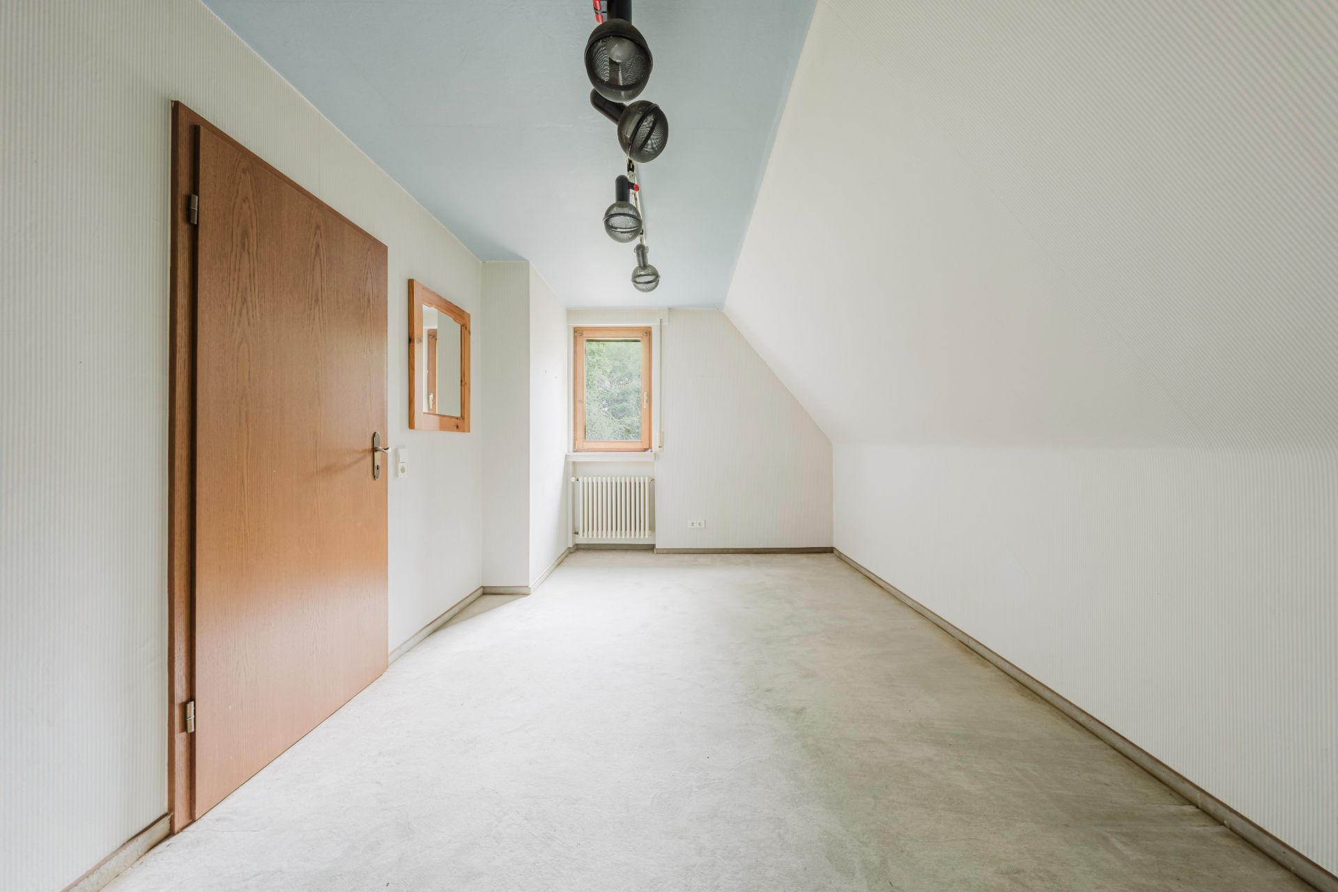 Leerer Raum mit schräger Decke, Holztür, Fenster, Heizkörper und Deckenleuchte.