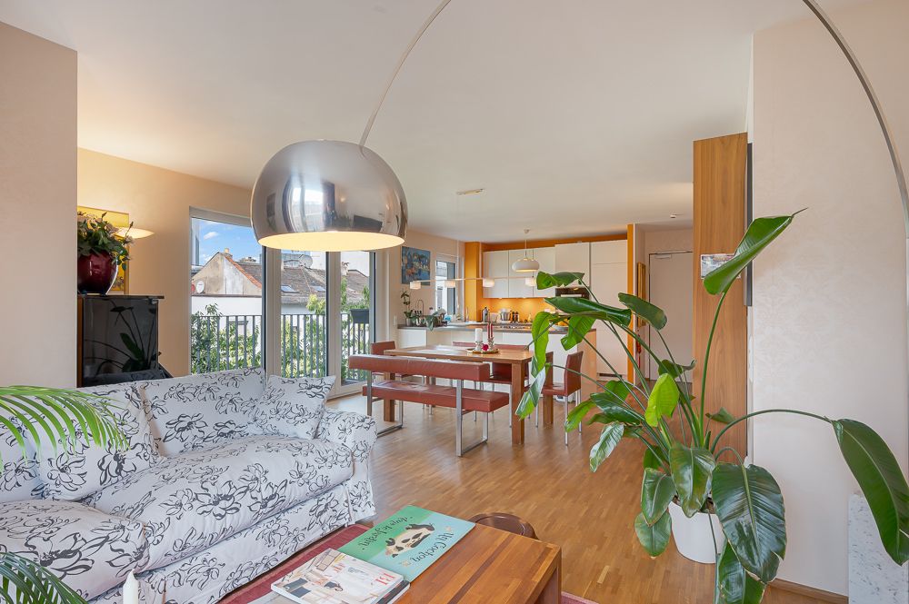 Helles Wohnzimmer mit gemusterter Couch, großer Hängelampe, Esstisch und Zimmerpflanze.