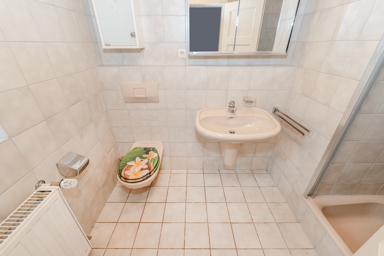 Kleines Badezimmer mit Toilette, Waschbecken, Spiegel und Dusche, Toilettendeckel mit Blumenmotiv.