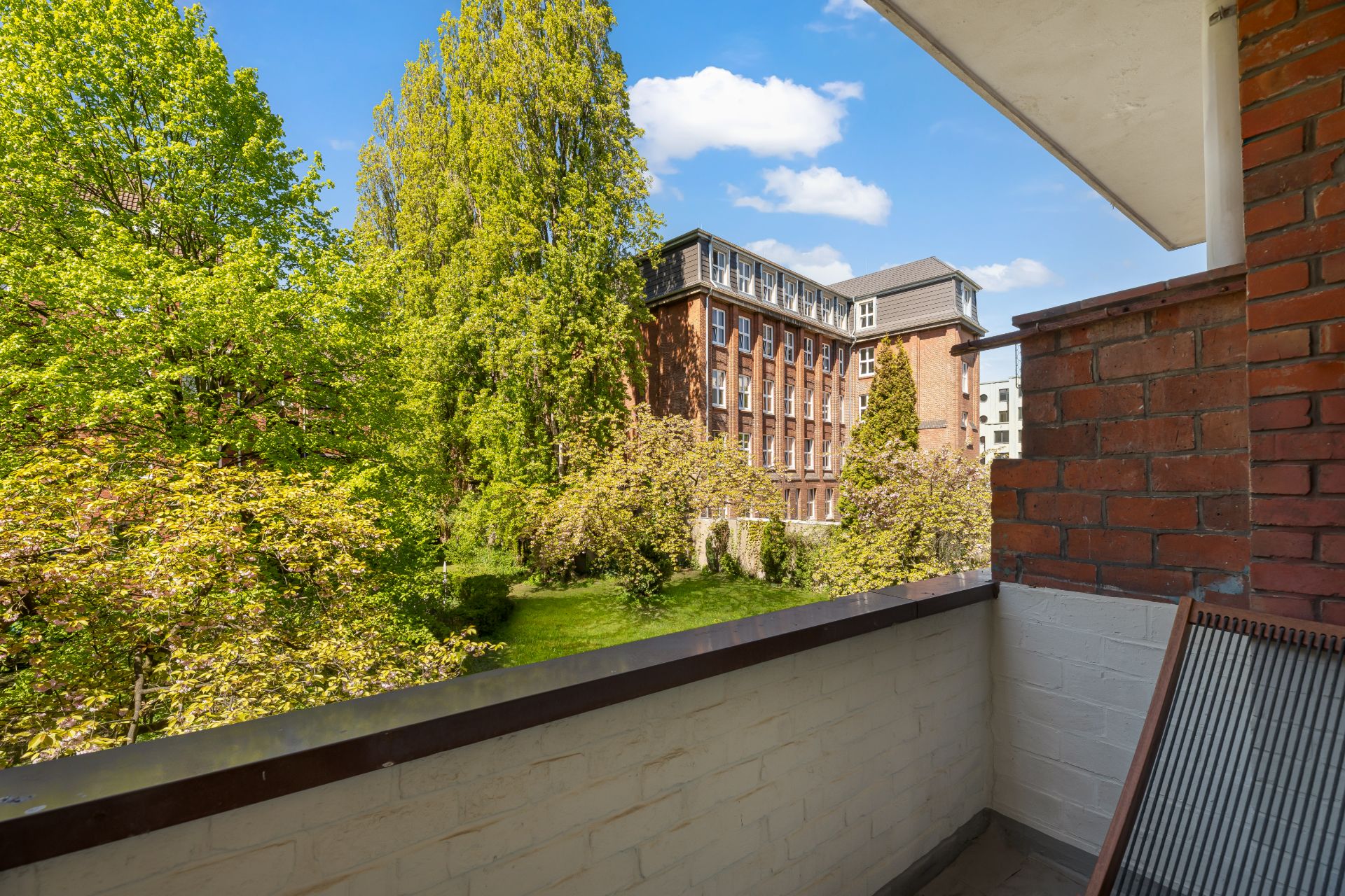 Blick von einem Balkon auf grüne Bäume und ein mehrstöckiges Backsteingebäude.