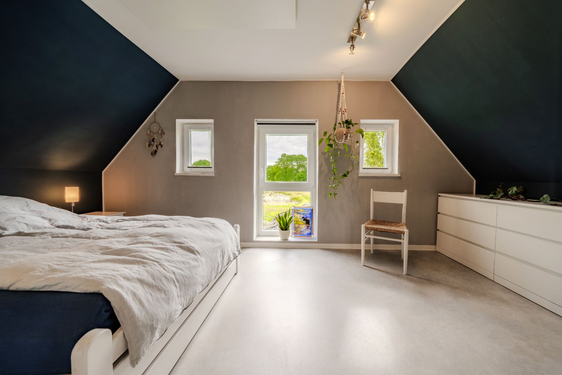 Schlafzimmer mit großem Bett, Kommode, Stuhl und drei Fenstern mit Blick ins Grüne.