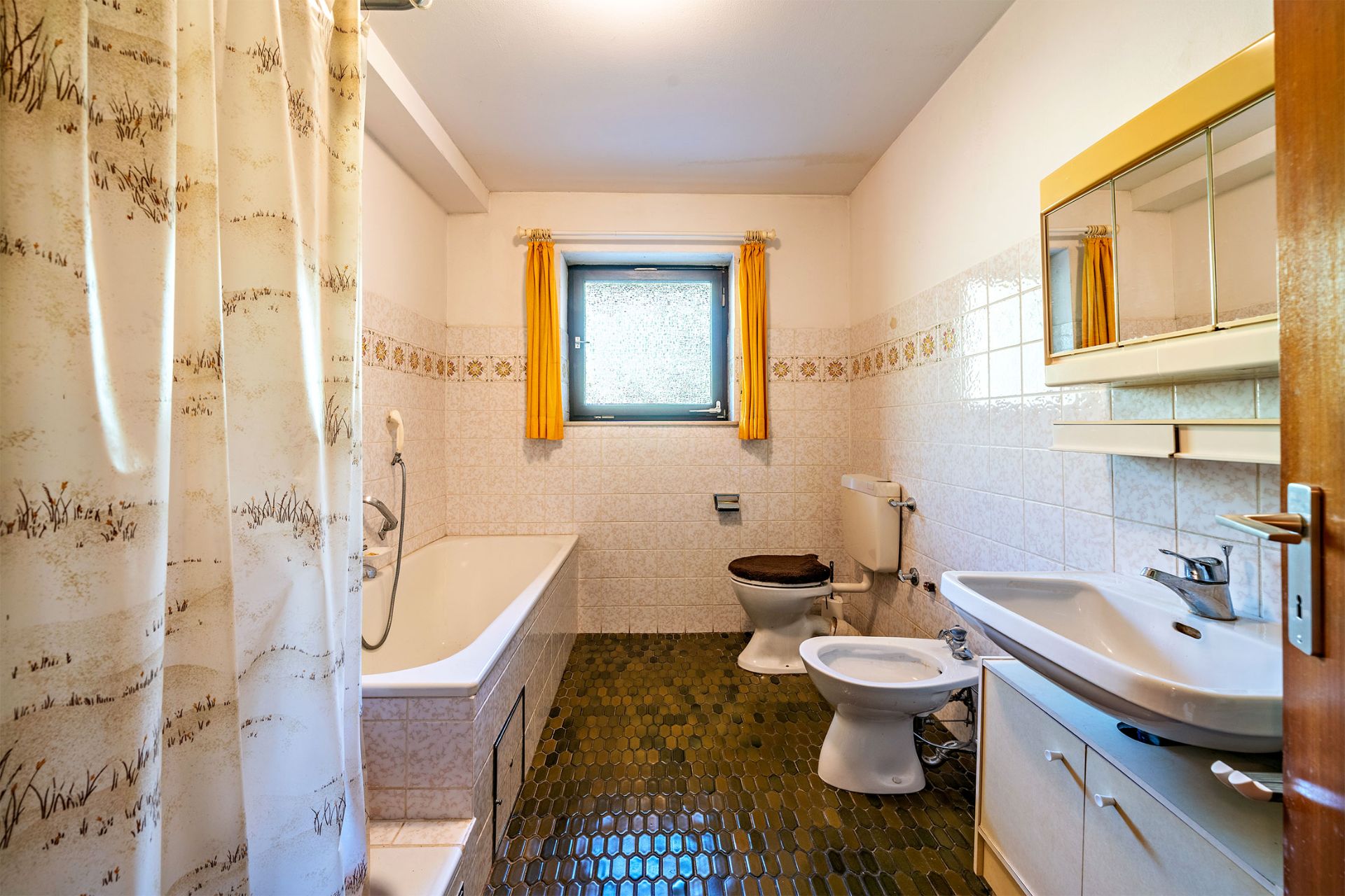 Badezimmer mit Badewanne, Toilette, Waschbecken, Spiegel und Fenster mit gelben Vorhängen.