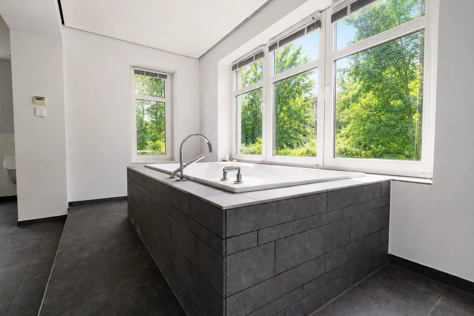 Modernes Badezimmer mit großer Badewanne vor drei großen Fenstern und grauen Fliesen.