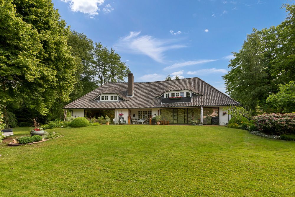 Großes Einfamilienhaus mit Garten, Bäumen und Rasenfläche bei blauem Himmel.