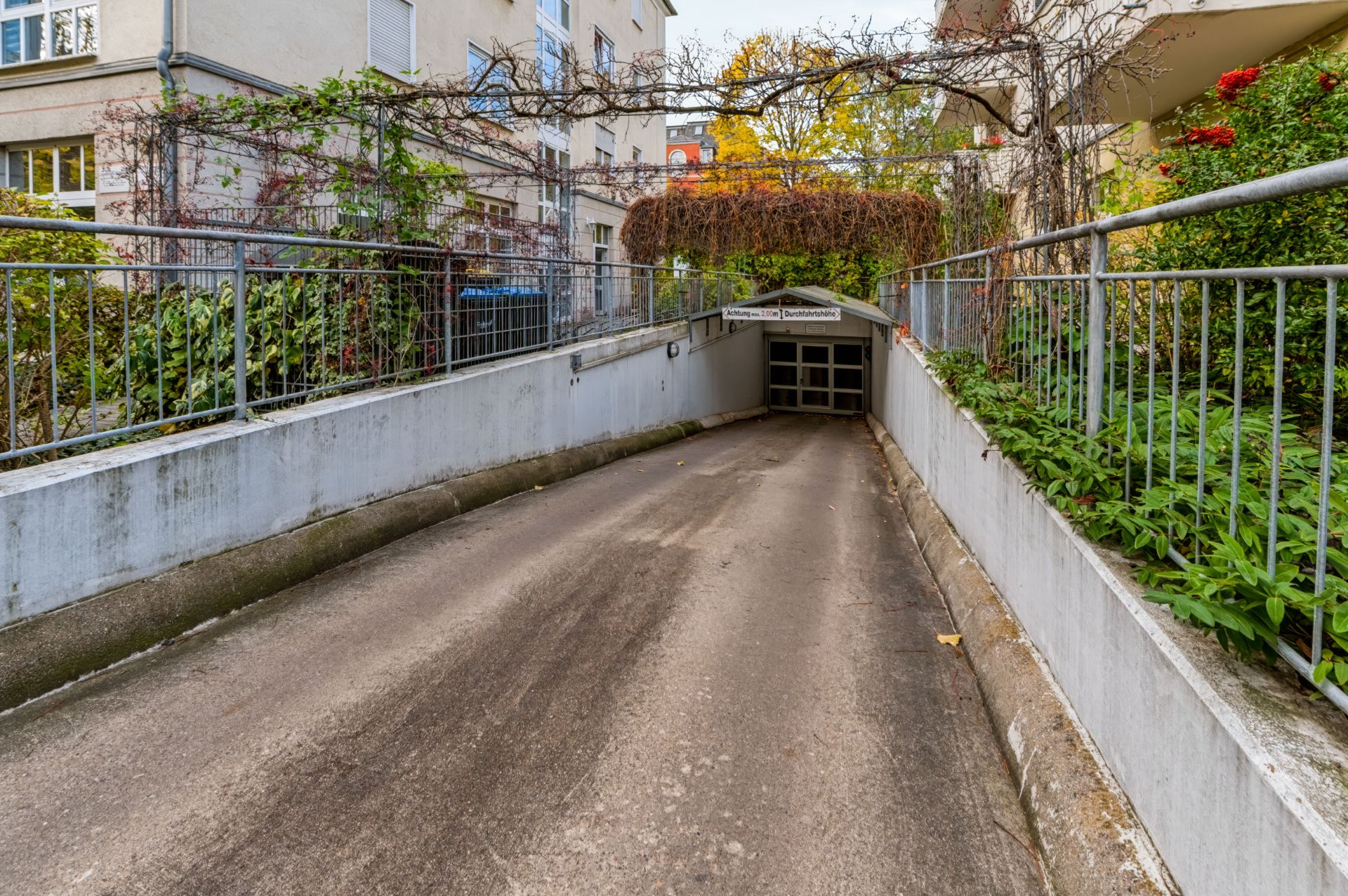 Einfahrt zu einer Tiefgarage mit Pflanzen an den Seiten und Wohngebäuden im Hintergrund.