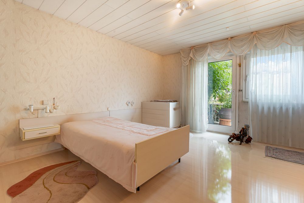Helles Schlafzimmer mit Doppelbett, Kommode, Teppich und Zugang zu einem Balkon mit viel Grün.