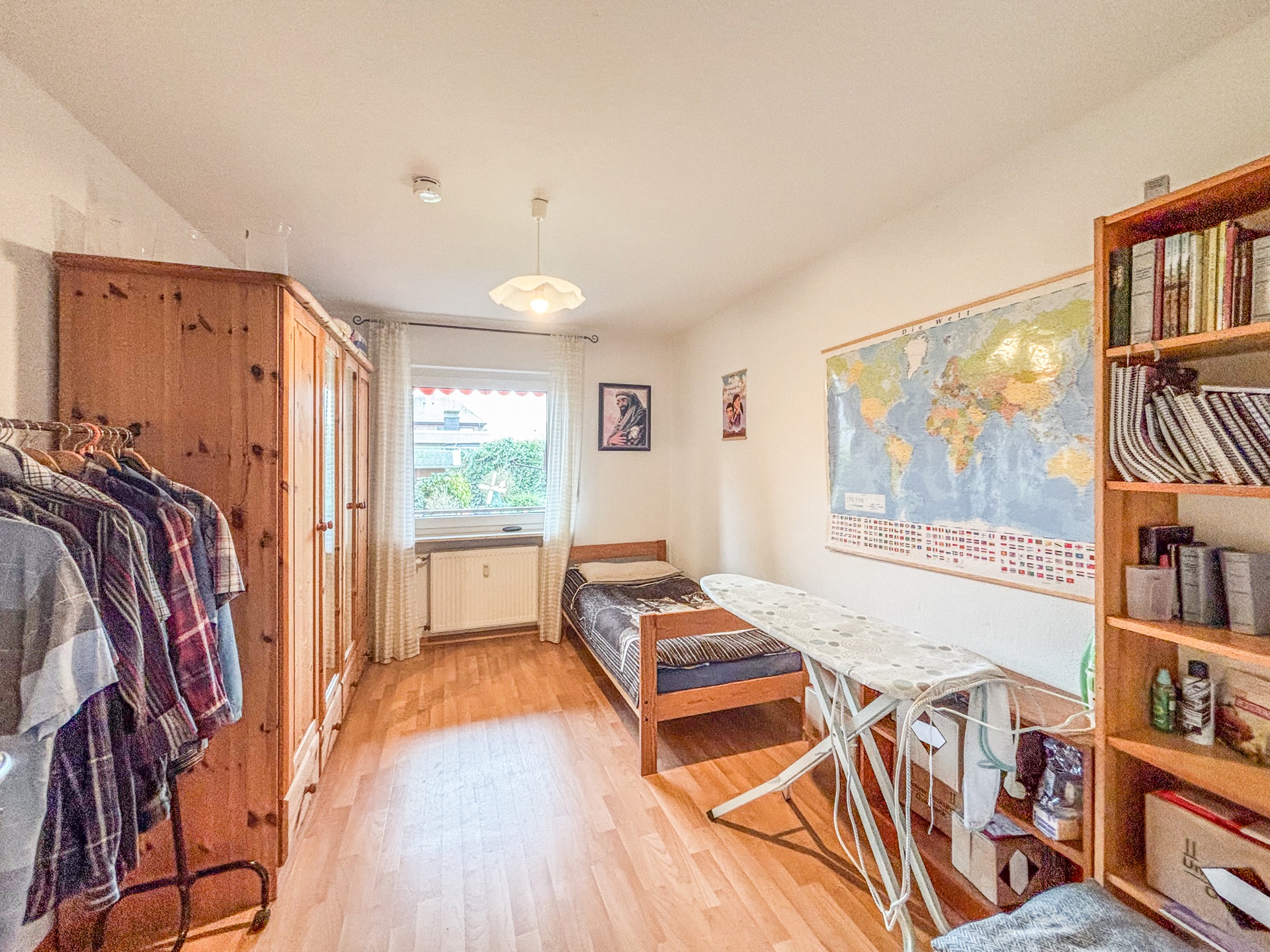 Helles Schlafzimmer mit Einzelbett, Schrank, Bücherregal, Kleiderständer und Bügelbrett.