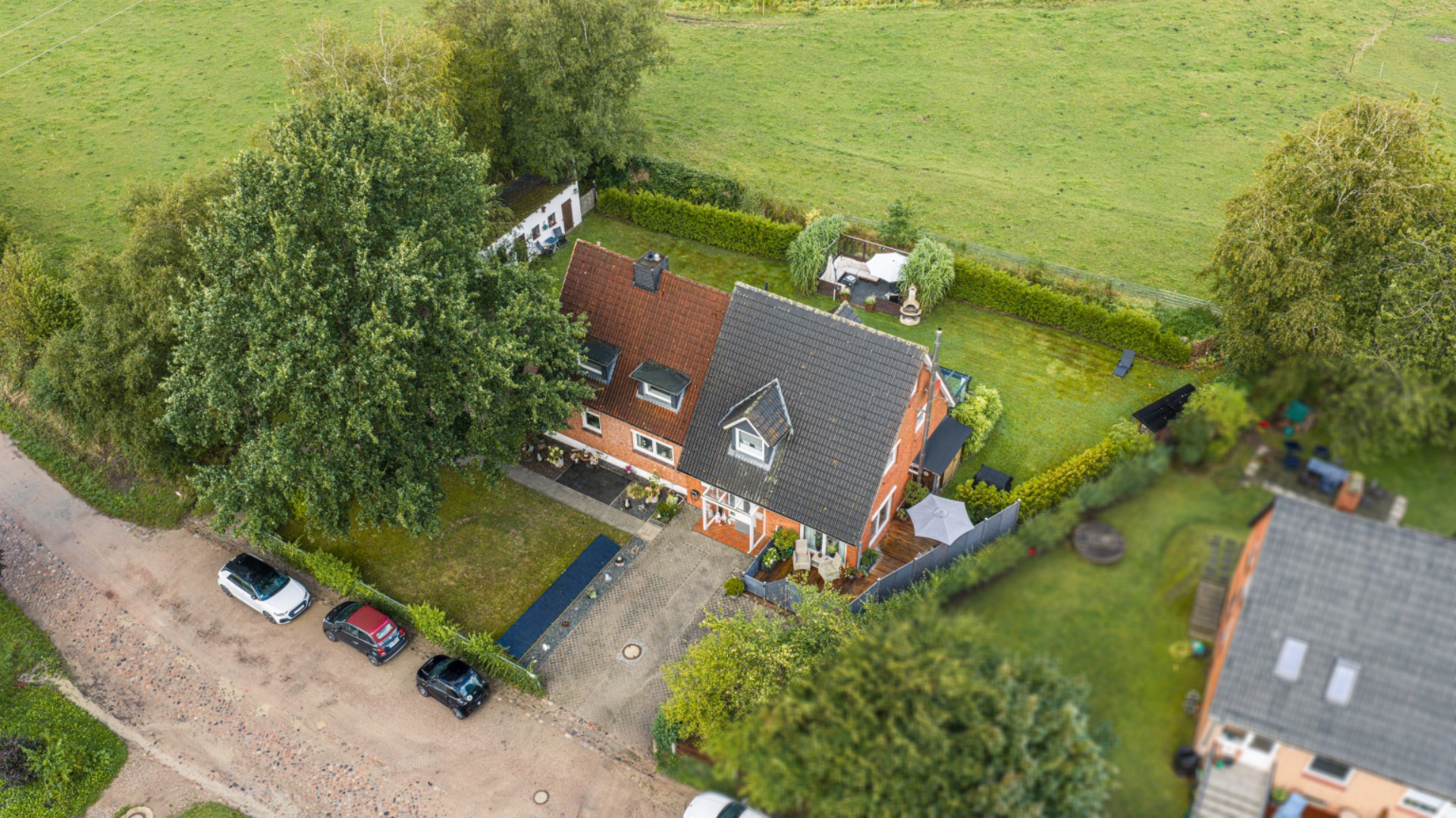 Luftaufnahme eines Backsteinhauses mit Garten und angrenzender Wiese, mehrere Autos parken davor.