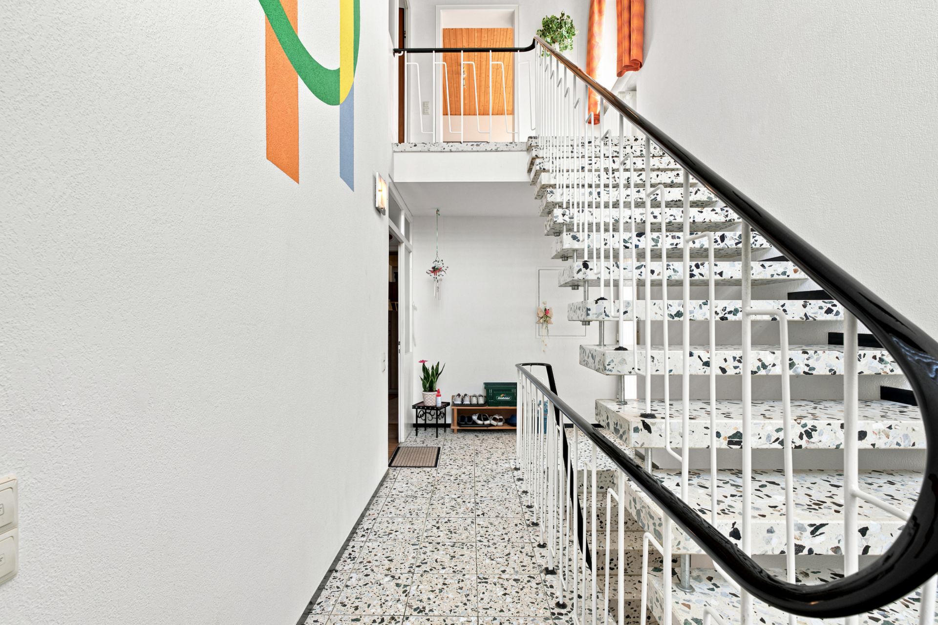 Heller Hausflur mit Treppe, Terrazzo-Boden und bunter Wandmalerei.