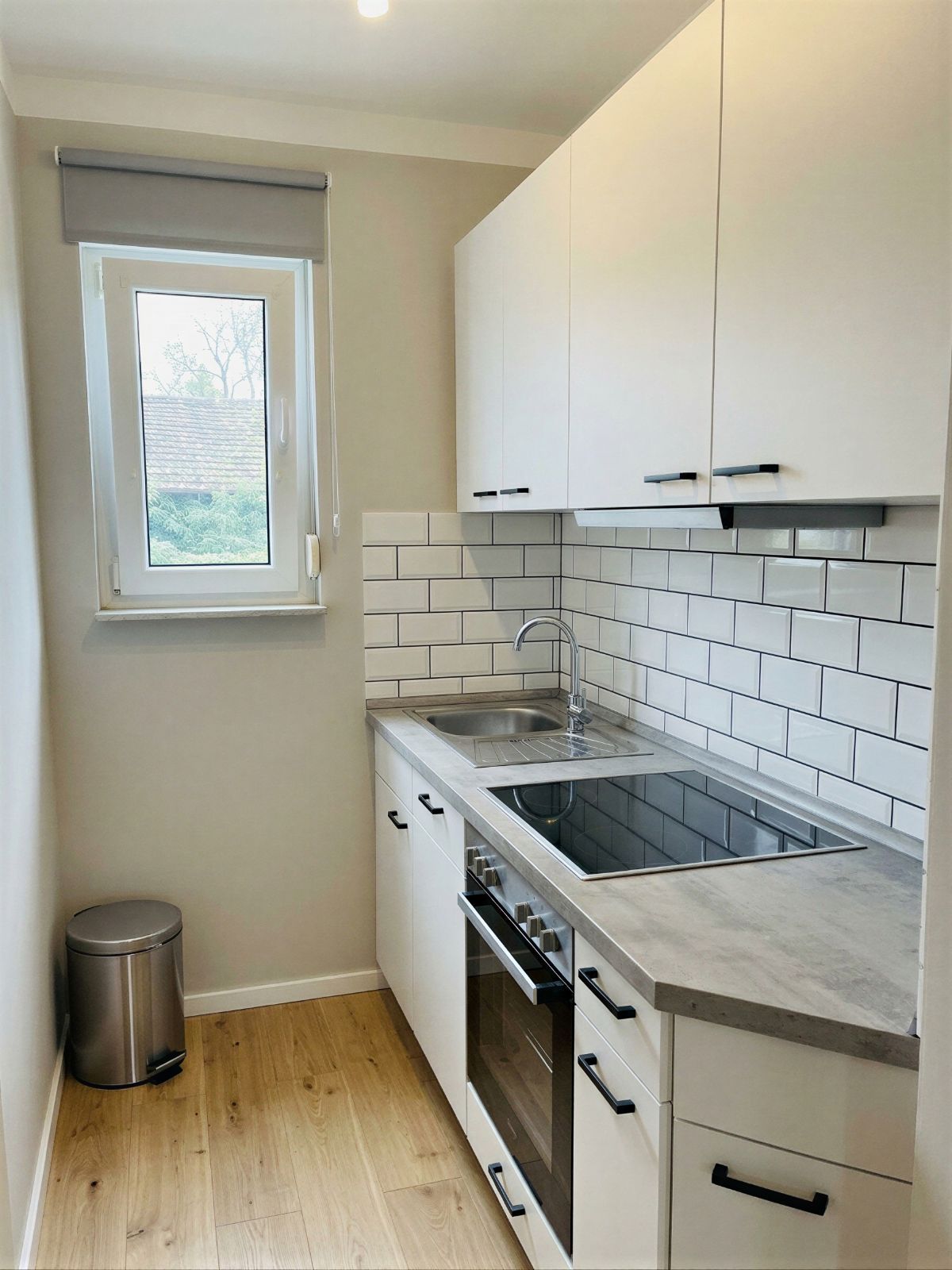 Schmale Küche mit weißen Schränken, Herd, Spüle, Fenster und Mülleimer auf Holzfußboden.