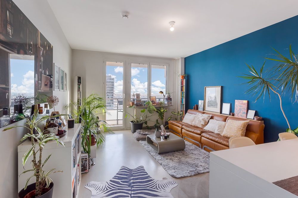 Modernes Wohnzimmer mit braunem Sofa, blauer Wand, Pflanzen und großen Fenstern mit Blick nach draußen.