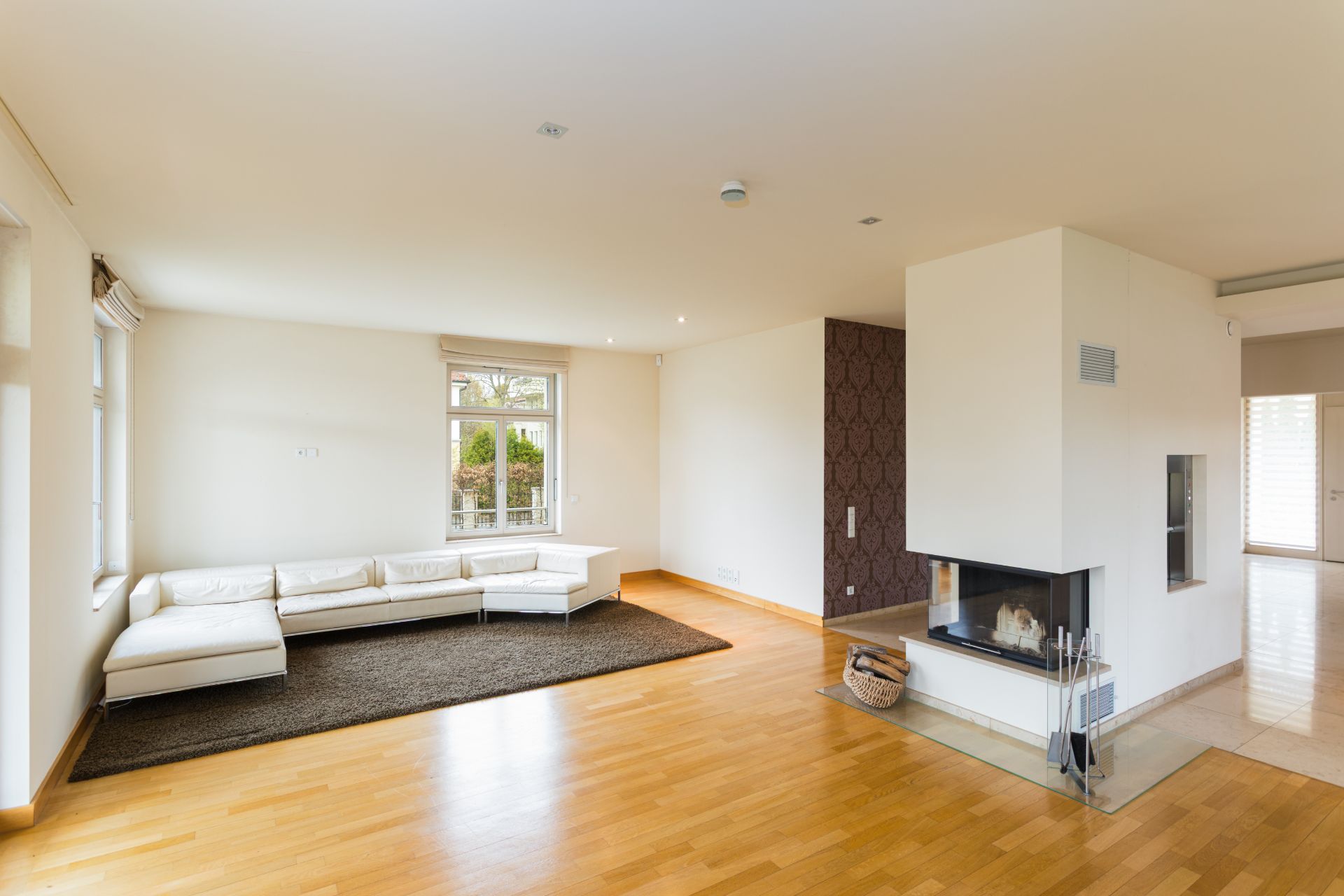 Helles Wohnzimmer mit Ecksofa, Kamin und großem Fenster, Holzboden und Teppich.