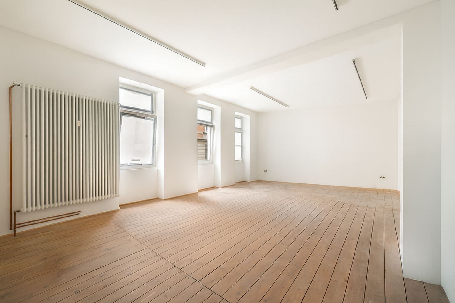 Leerer, heller Raum mit Holzboden, weißen Wänden, drei Fenstern und einem großen Heizkörper.
