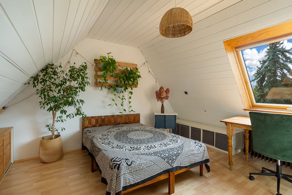 Schlafzimmer mit Bett, Schreibtisch am Fenster, Pflanzen und Holzdecke.