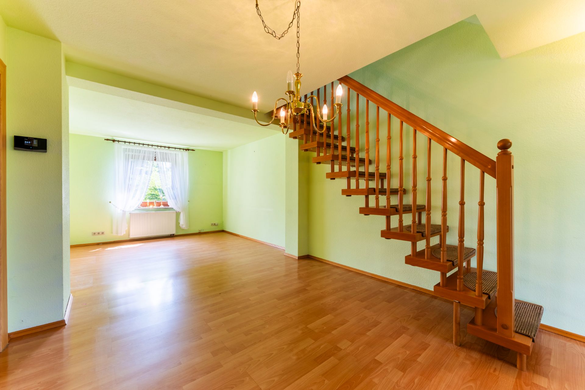 Helles Wohnzimmer mit Holztreppe, Parkettboden und Fenster mit weißen Vorhängen.