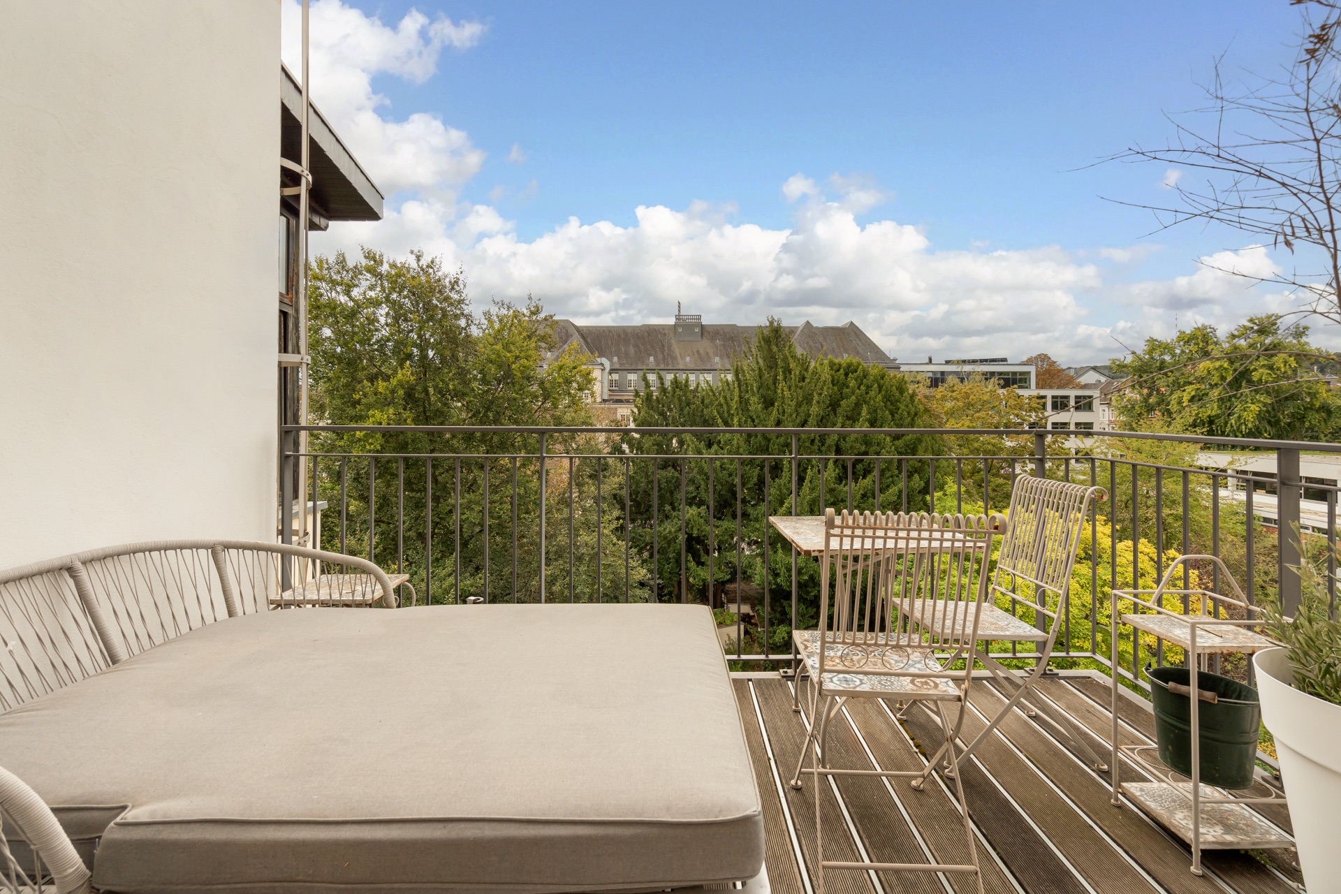 Balkon mit Sitzbank, Metalltisch und Stühlen, Blick auf Bäume und Gebäude im Hintergrund.