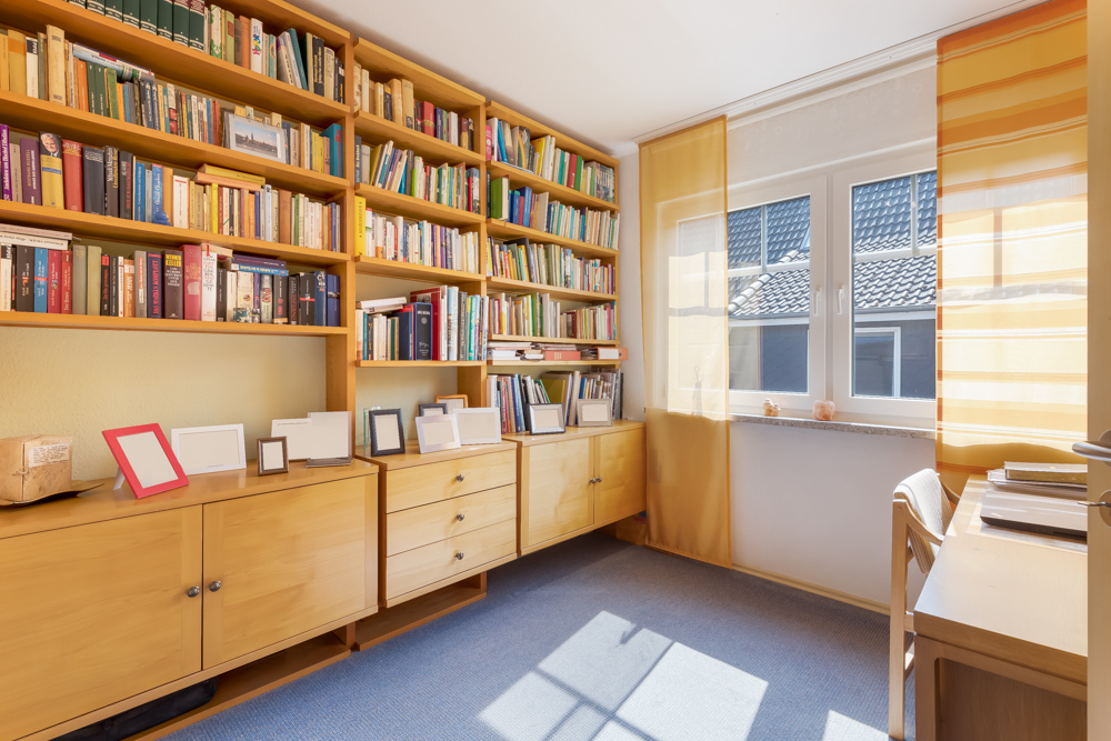Helles Arbeitszimmer mit Bücherregal, Schreibtisch und großem Fenster mit gelben Vorhängen.