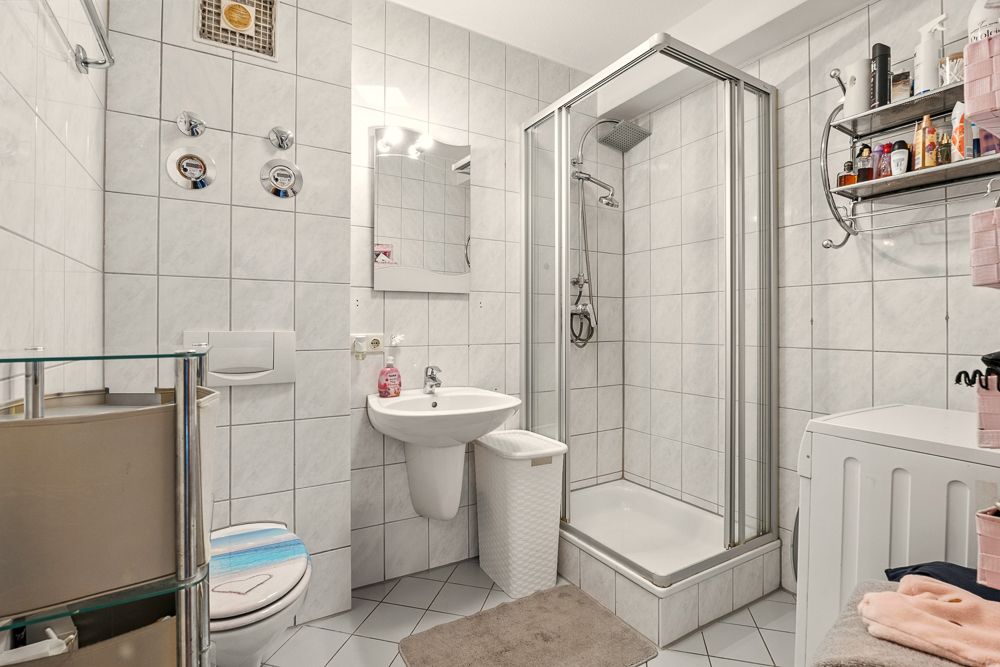 Helles Badezimmer mit Dusche, Waschbecken, WC, Waschmaschine und Regal, alles weiß gefliest.