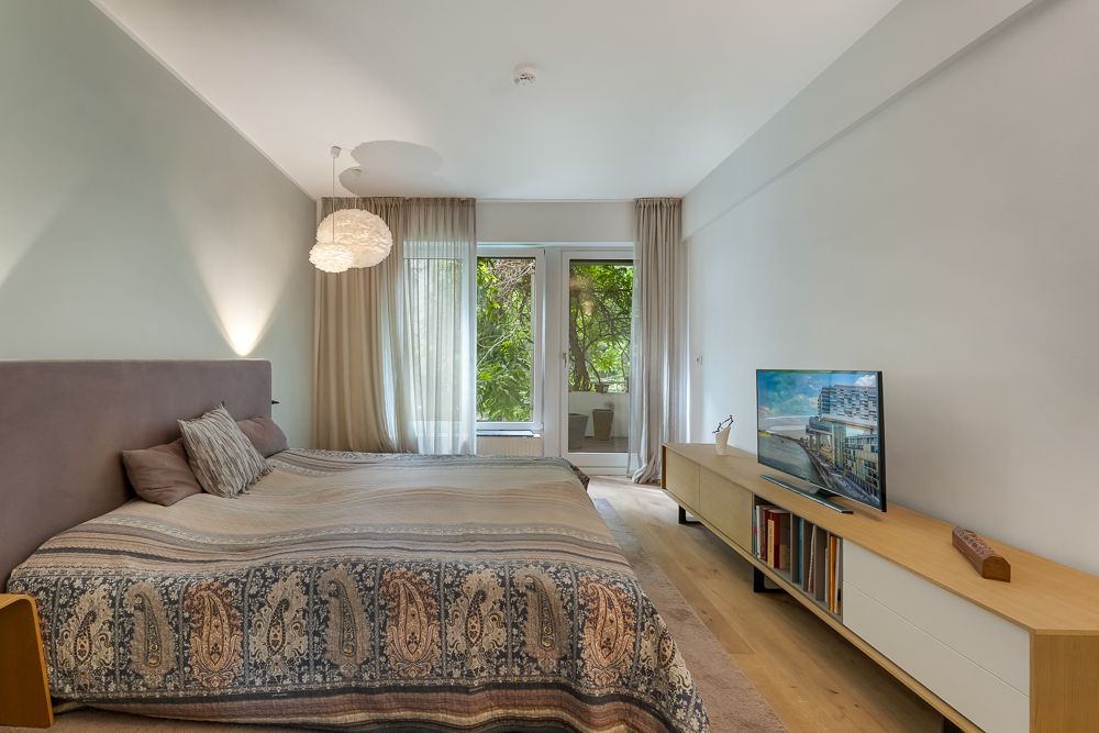 Schlafzimmer mit Doppelbett, Holzfußboden, Kommode mit Fernseher und Zugang zum Balkon.