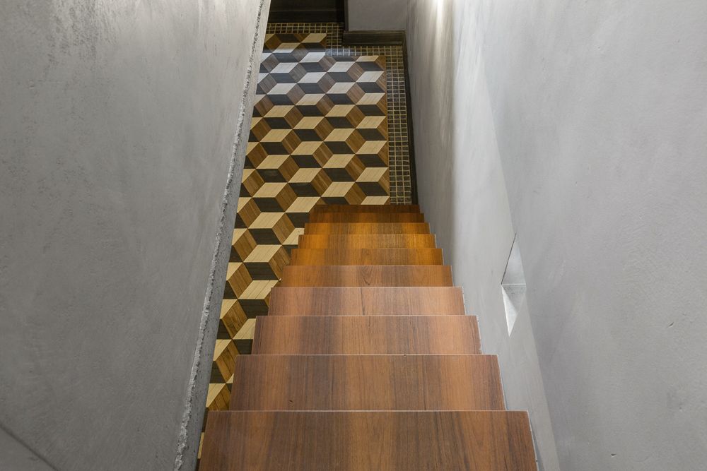 Holztreppe mit geometrisch gemustertem Fliesenboden am unteren Ende zwischen grauen Wänden.
