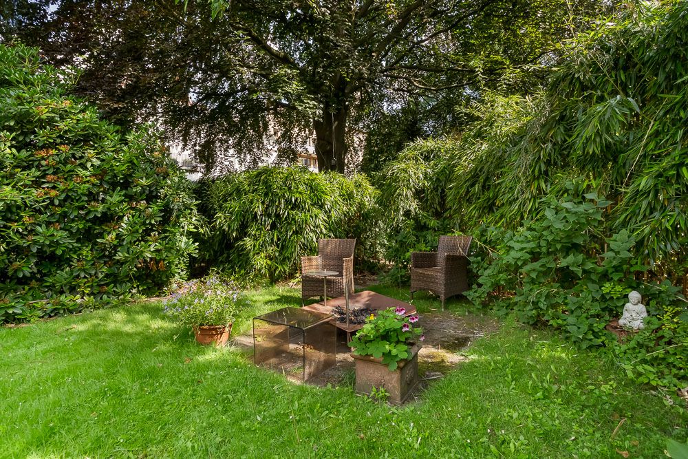 Garten mit zwei Korbstühlen, kleinem Tisch und viel grünem Bewuchs umgeben.