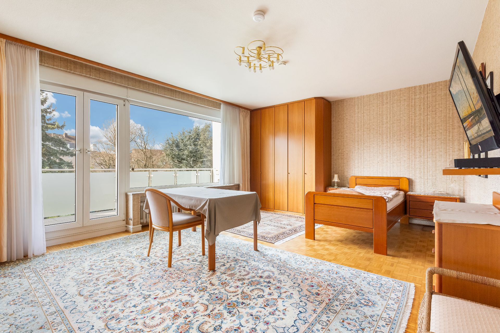 Helles Schlafzimmer mit Einzelbett, großem Teppich, Tisch und Schrank. Zugang zum Balkon durch Glastür.