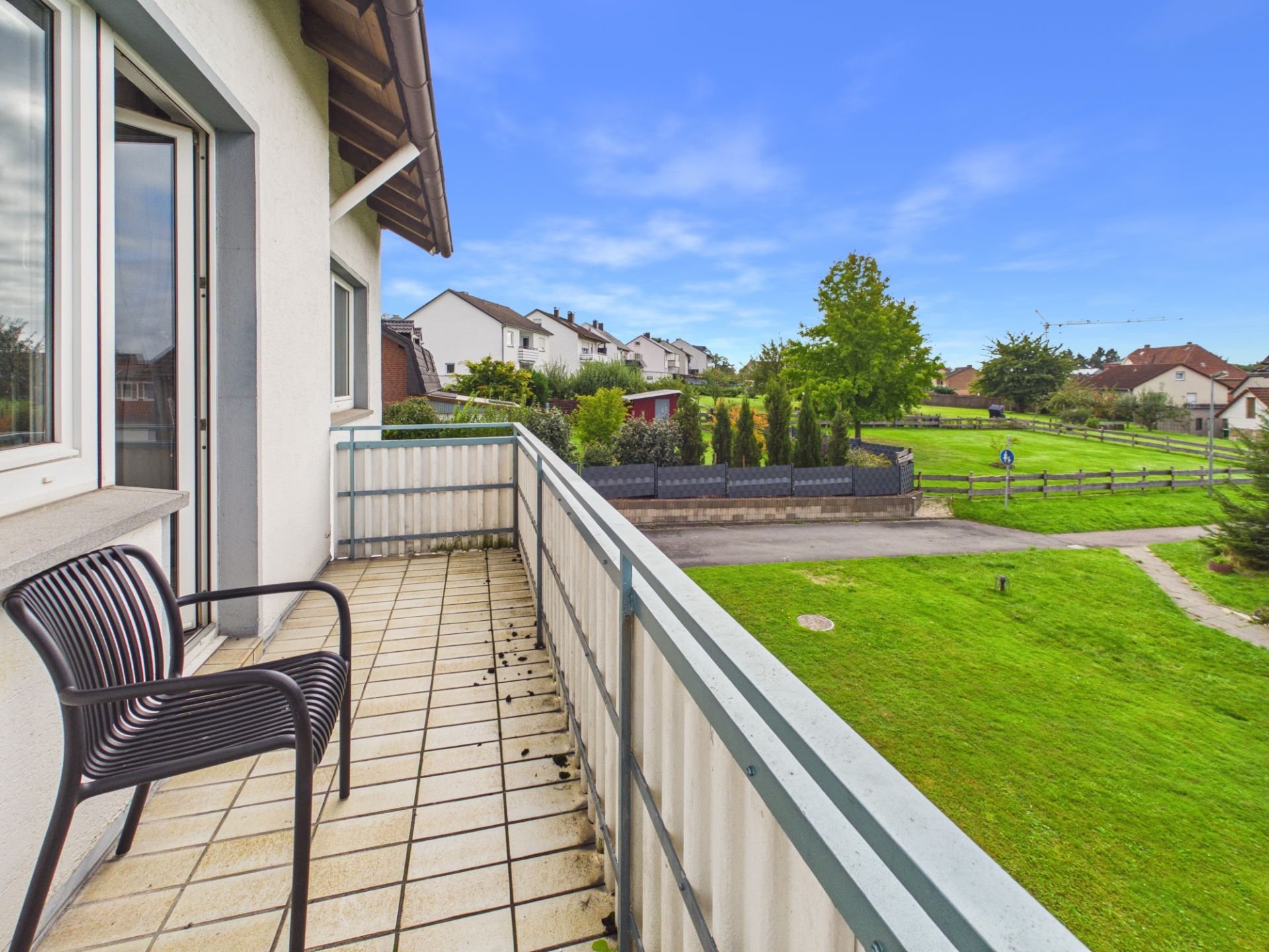 Balkon mit Stuhl, Fliesenboden und Ausblick auf Wiese, Weg und Wohnhäuser.