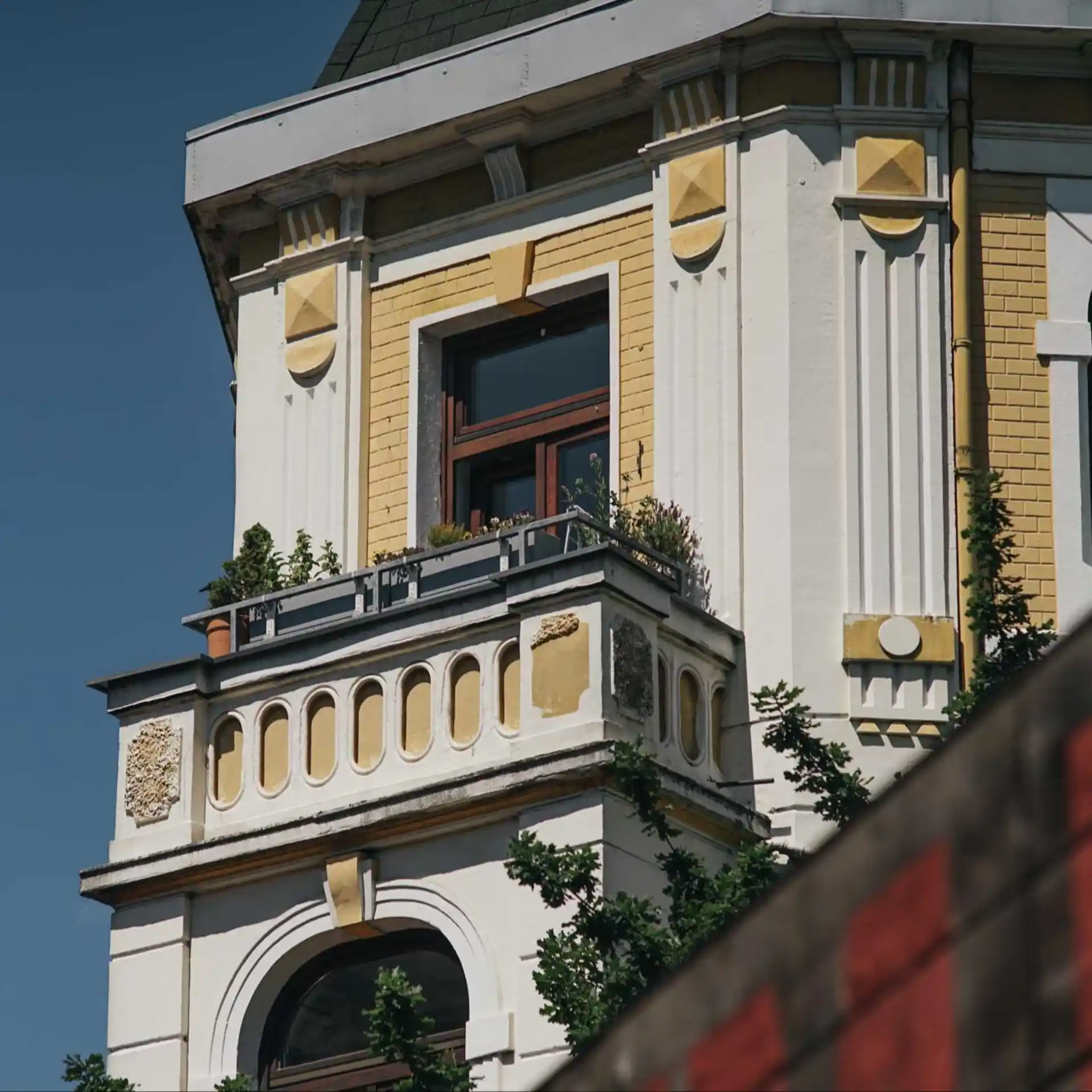 Balkon mit Pflanzen an einem alten Wohngebäude mit gelben und weißen Fassadenelementen.