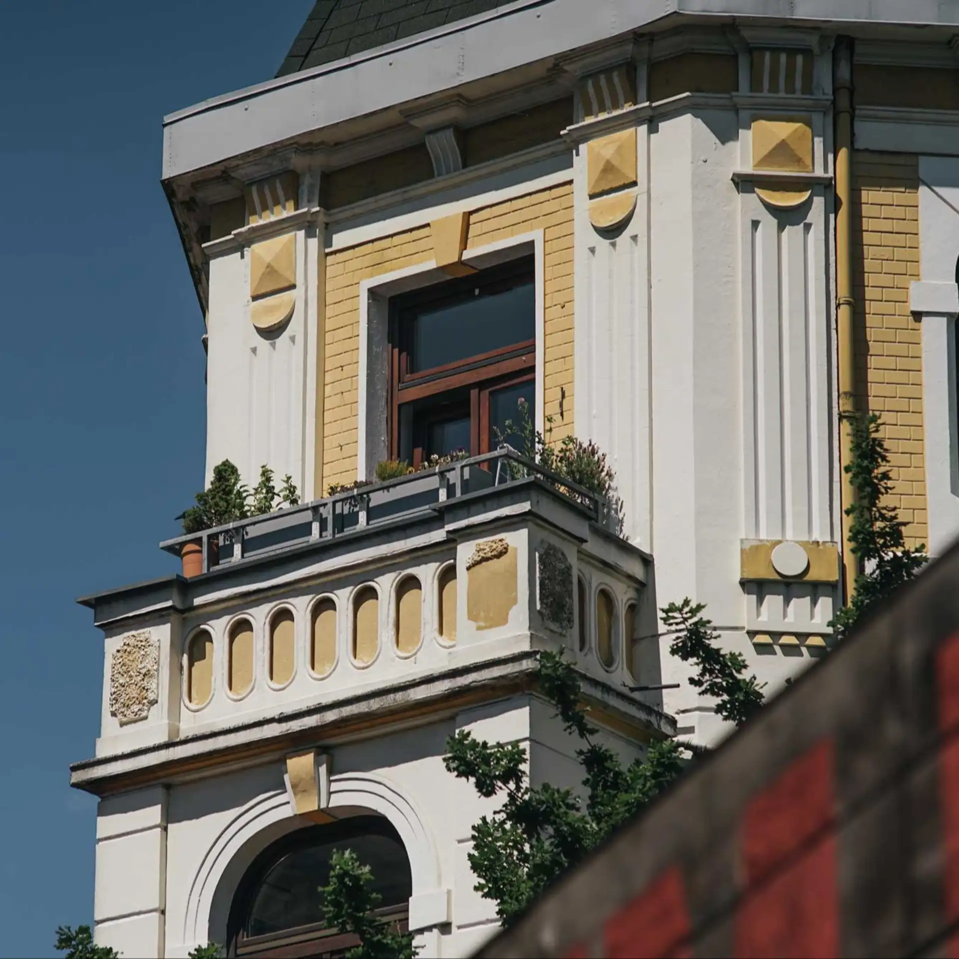 Balkon mit Pflanzen an einem alten Wohngebäude mit gelben und weißen Fassadenelementen.