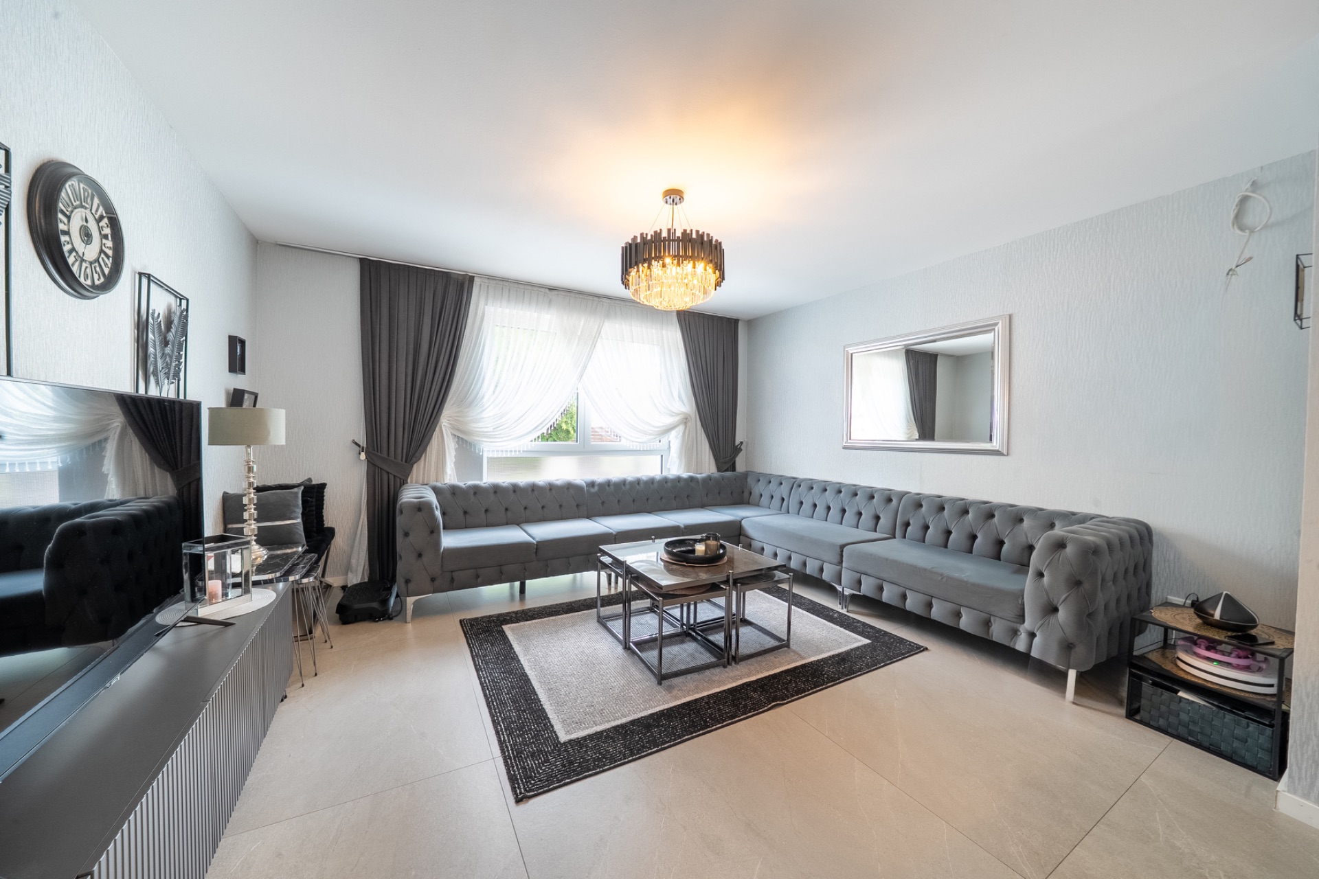 Wohnzimmer mit großem grauen Ecksofa, Couchtisch, Teppich, Spiegel und Kronleuchter.