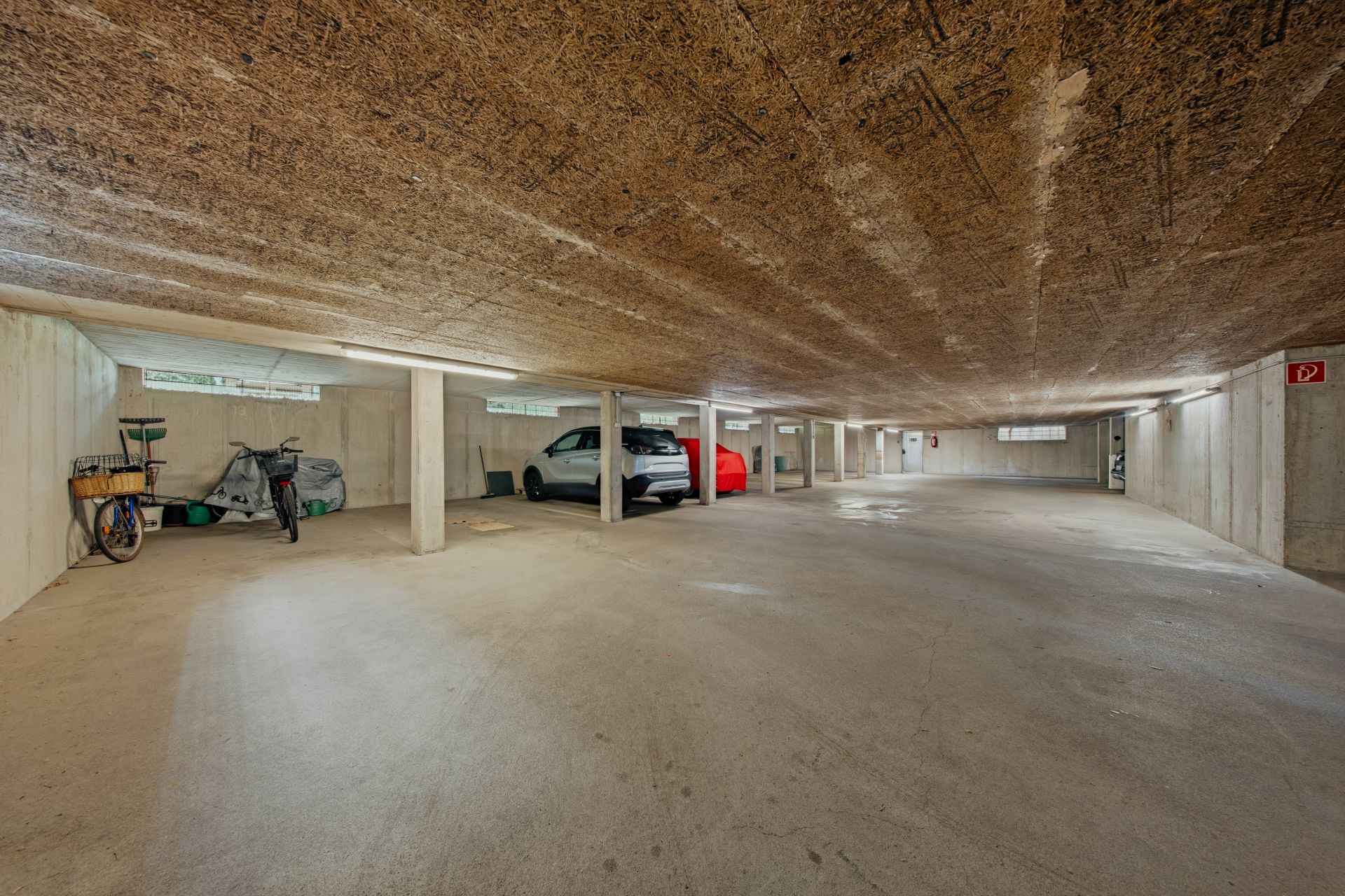 Tiefgarage mit geparkten Autos, Fahrrädern und Abstellflächen, Betonwände und niedrigem Holzdach.