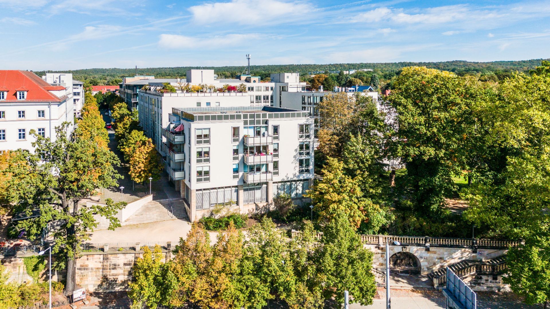 Modernes Wohngebäude mit Balkonen, umgeben von Bäumen und einer kleinen Mauer im Vordergrund.
