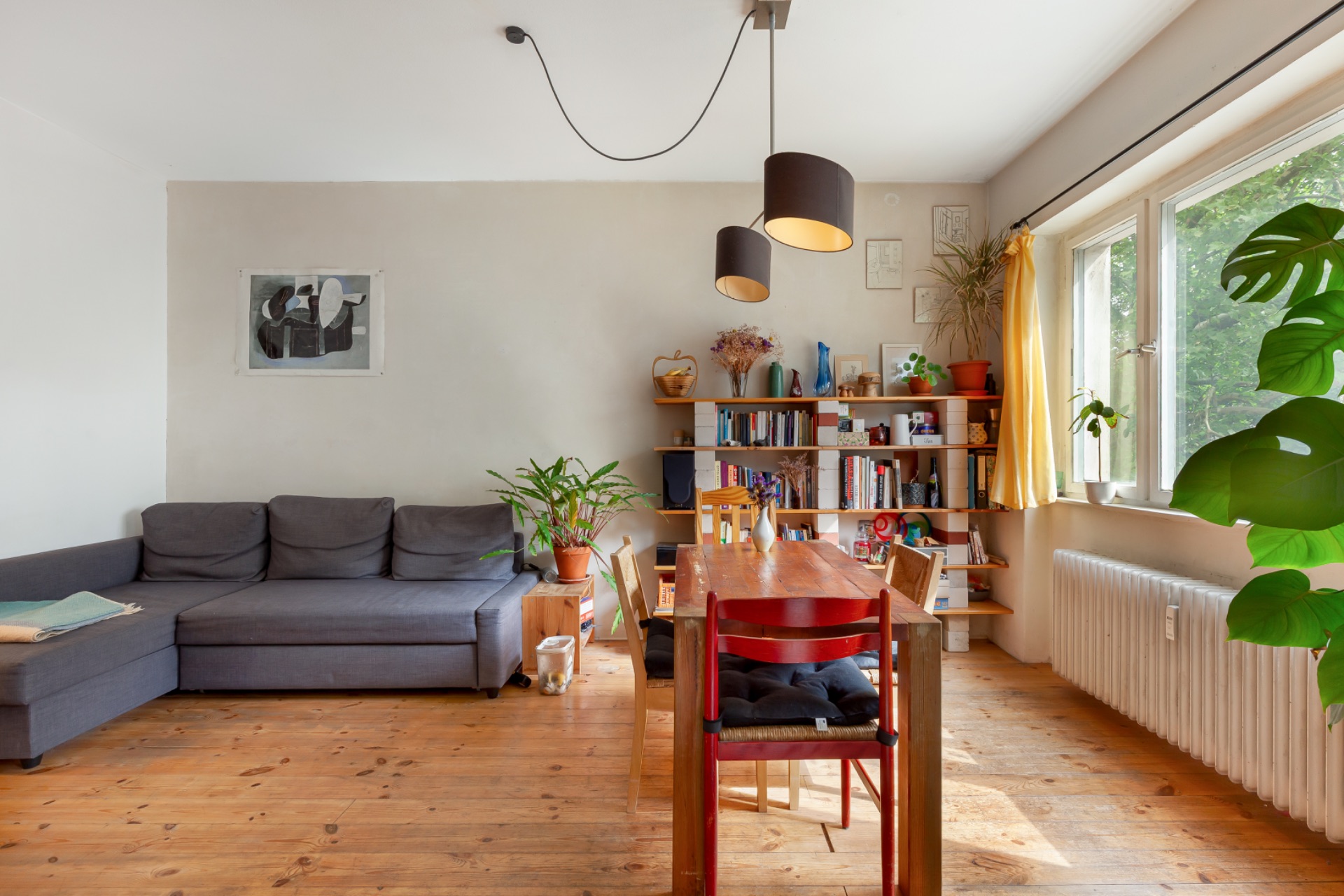 Helles Wohnzimmer mit grauem Sofa, Holztisch, Bücherregal und Pflanzen am Fenster.