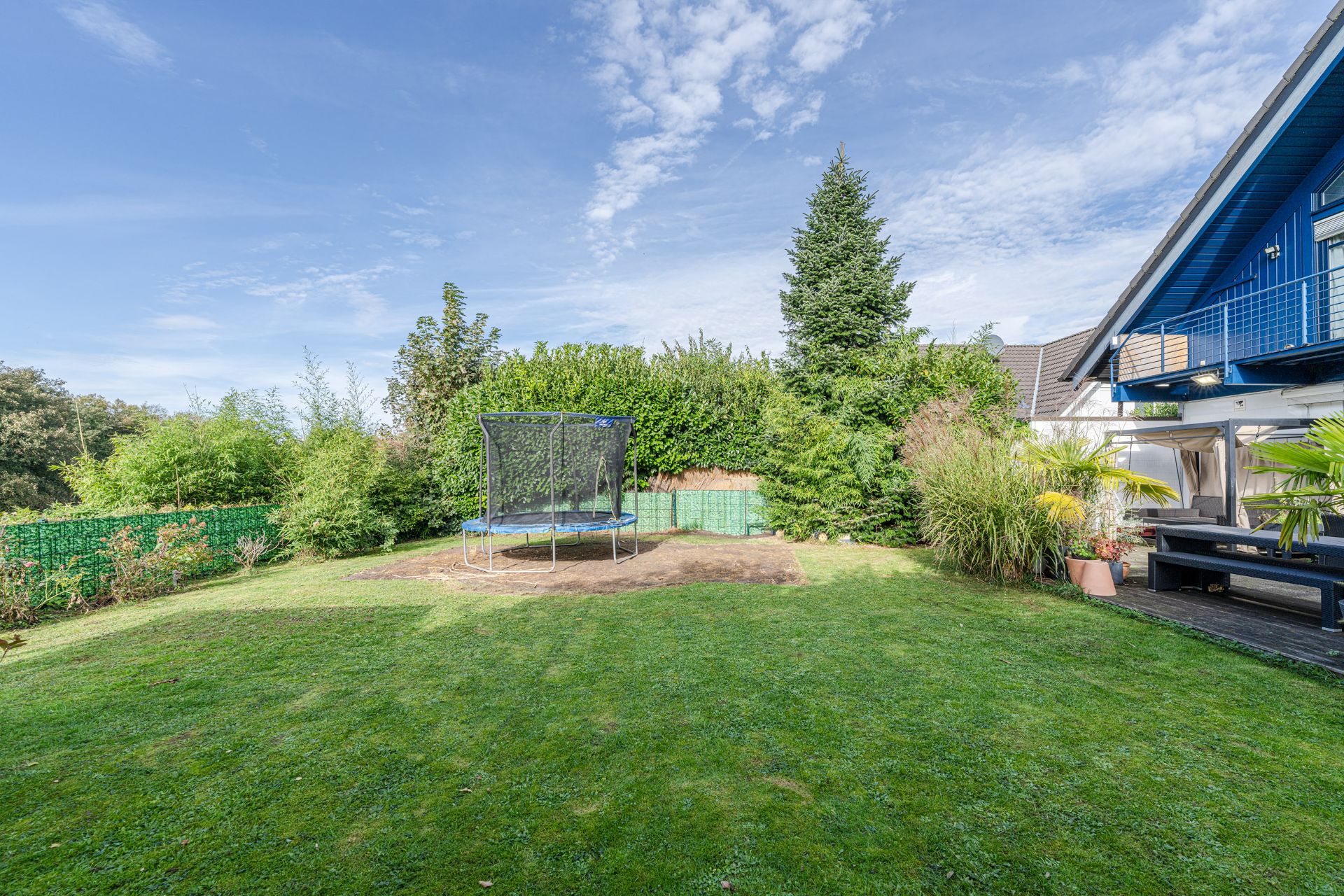 Grüner Garten mit Trampolin, umgeben von Sträuchern und einem Haus mit Terrasse.