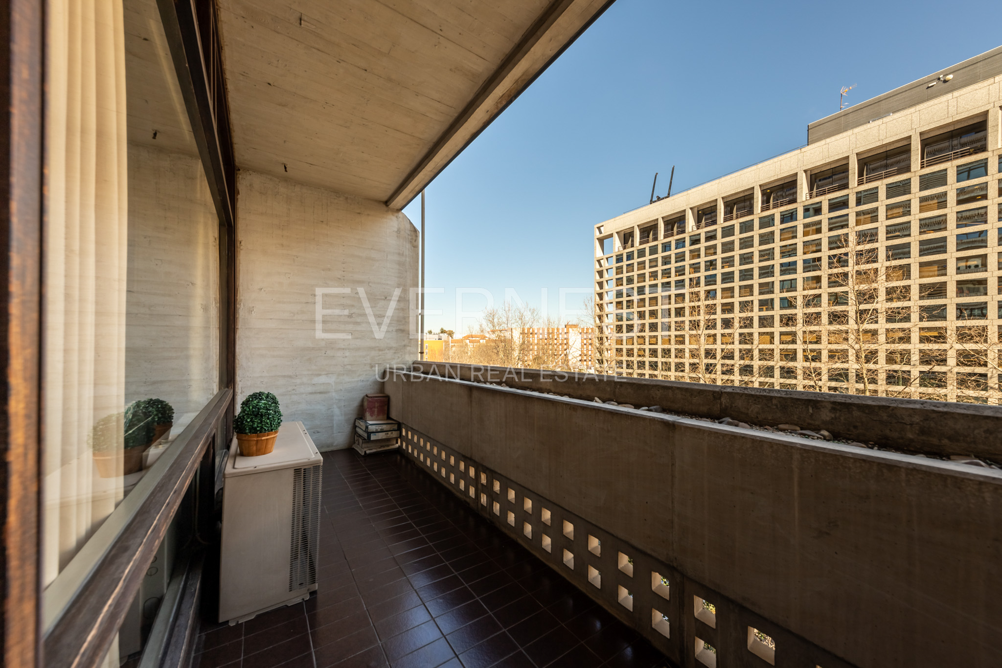 Überdachter Balkon mit braunen Fliesen, Topfpflanze und Blick auf ein großes Gebäude.