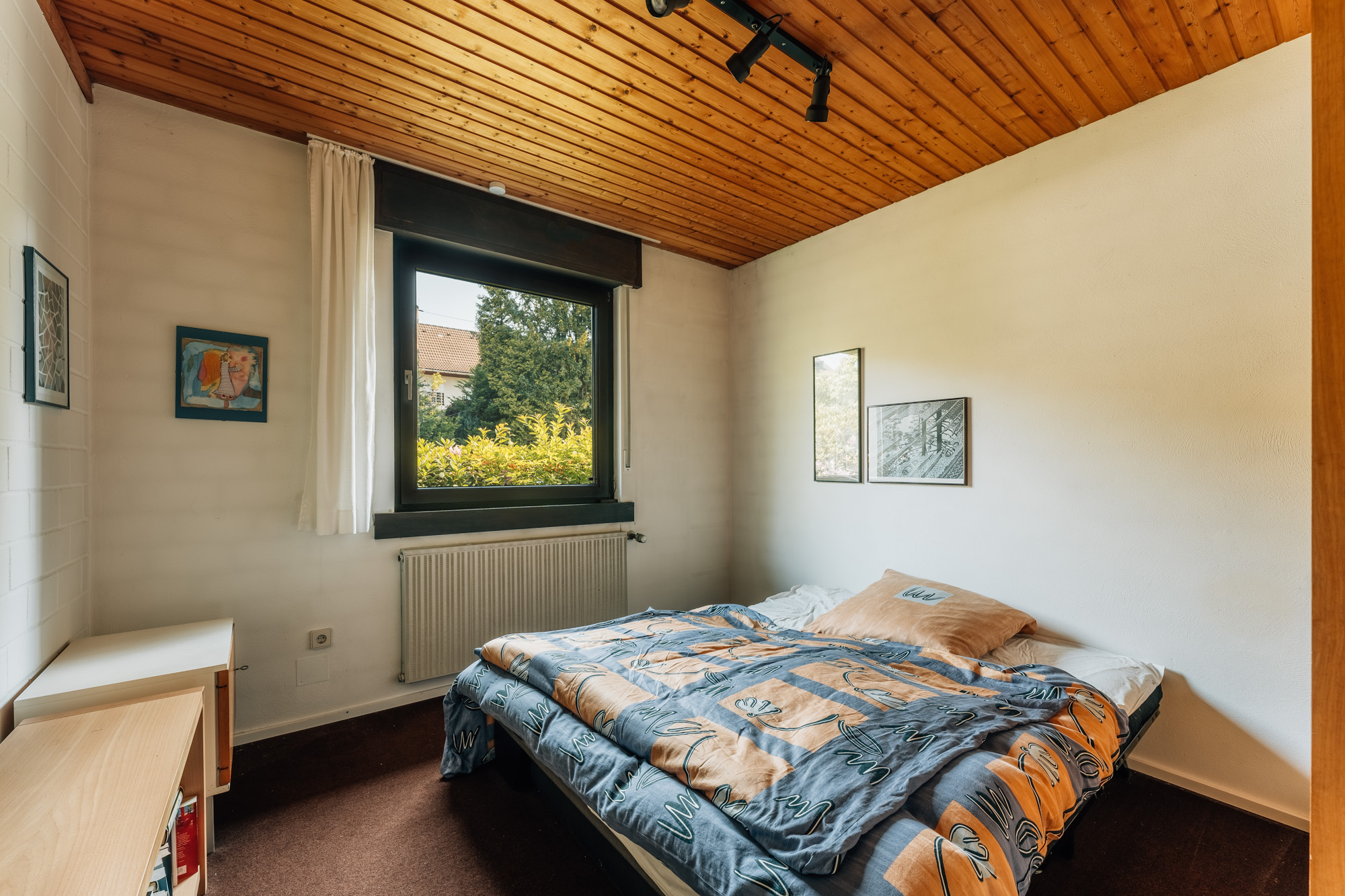 Kleines Schlafzimmer mit Holzbett, großem Fenster und Bildern an den weißen Wänden.