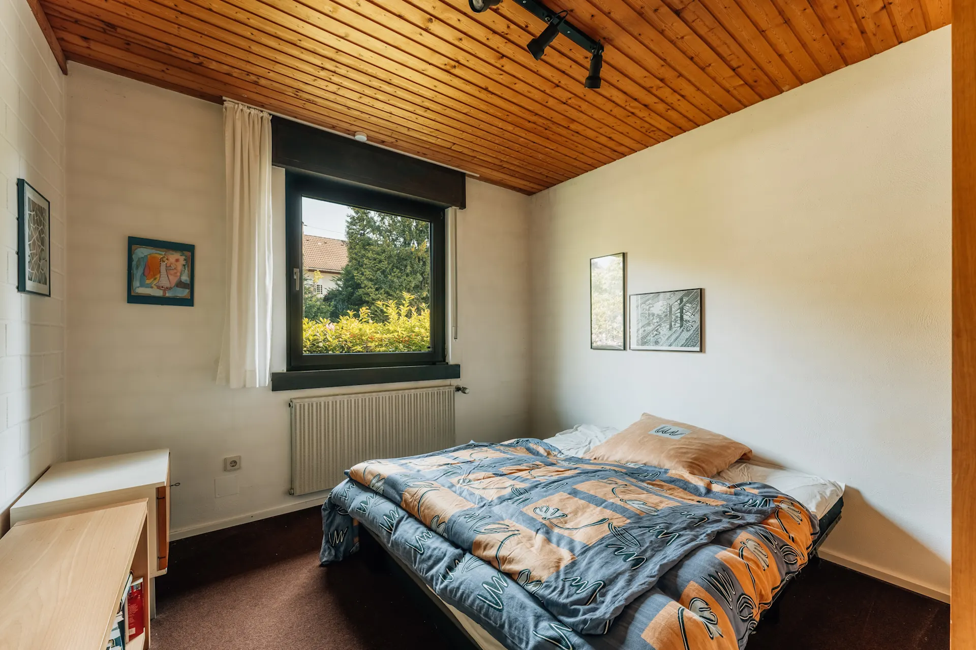 Kleines Schlafzimmer mit Holzbett, großem Fenster und Bildern an den weißen Wänden.