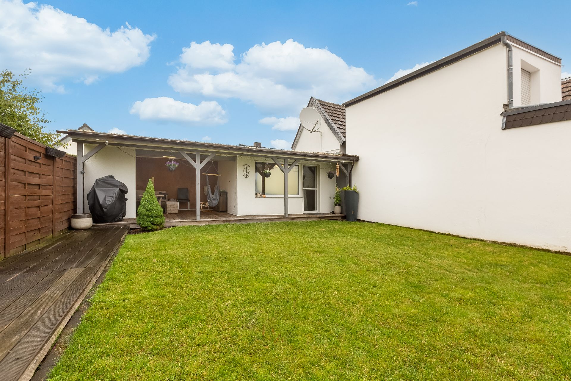 Garten mit Rasenfläche, Holzterrasse, überdachter Sitzbereich und weißen Hauswänden.