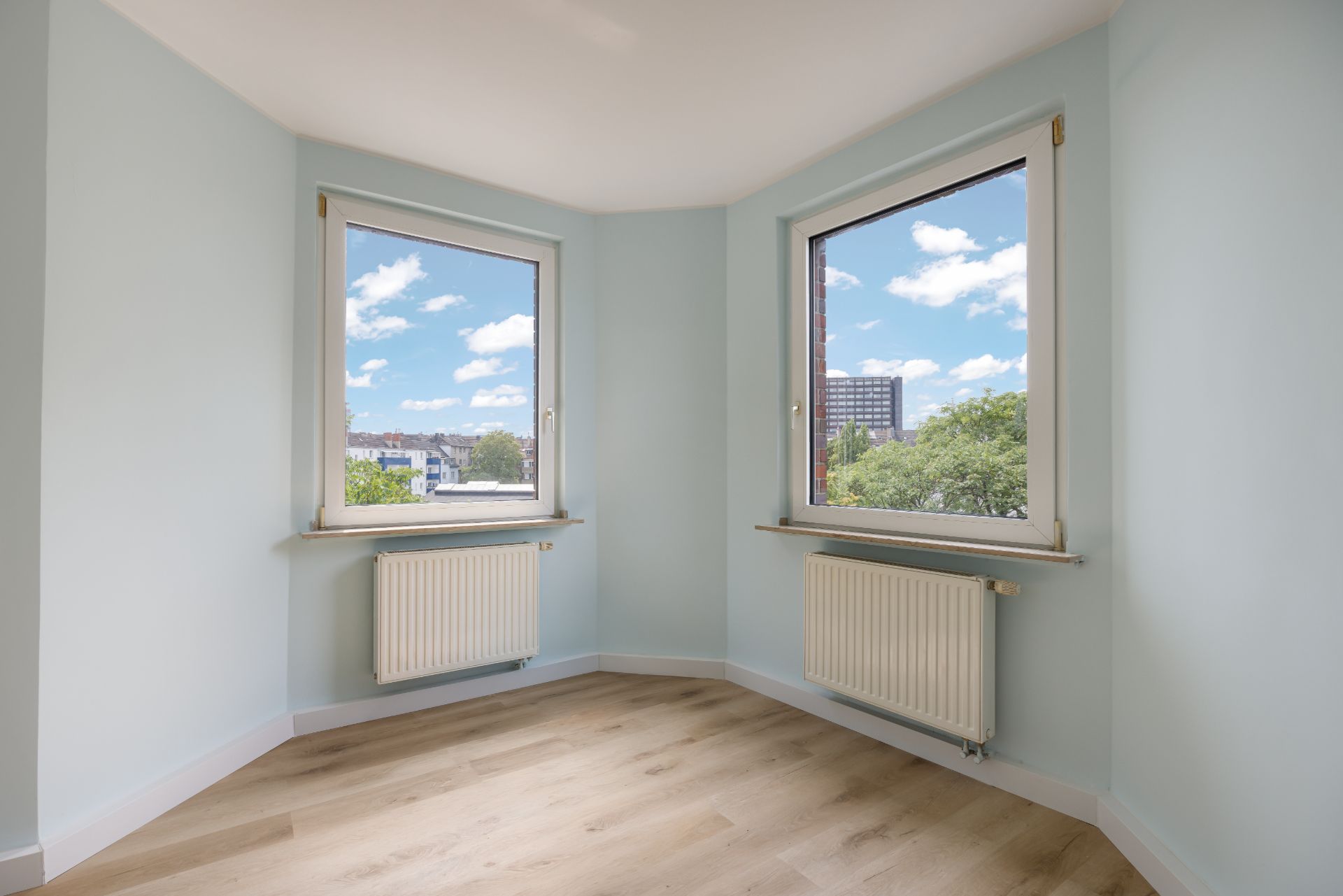 Helles Zimmer mit zwei Fenstern, Heizkörpern und hellem Holzfußboden.