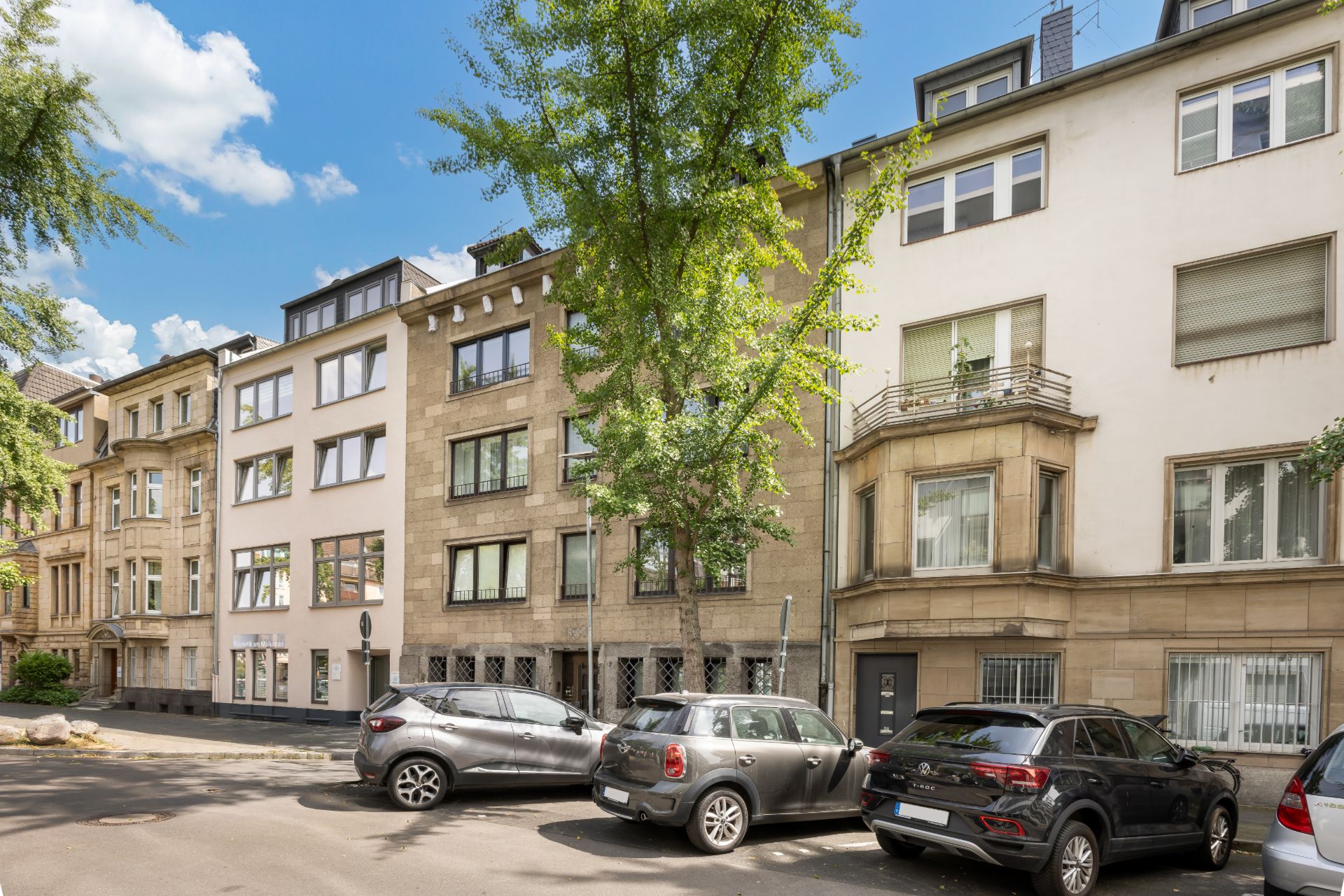 Mehrstöckige Wohnhäuser mit geparkten Autos und Bäumen an einer ruhigen Straße.