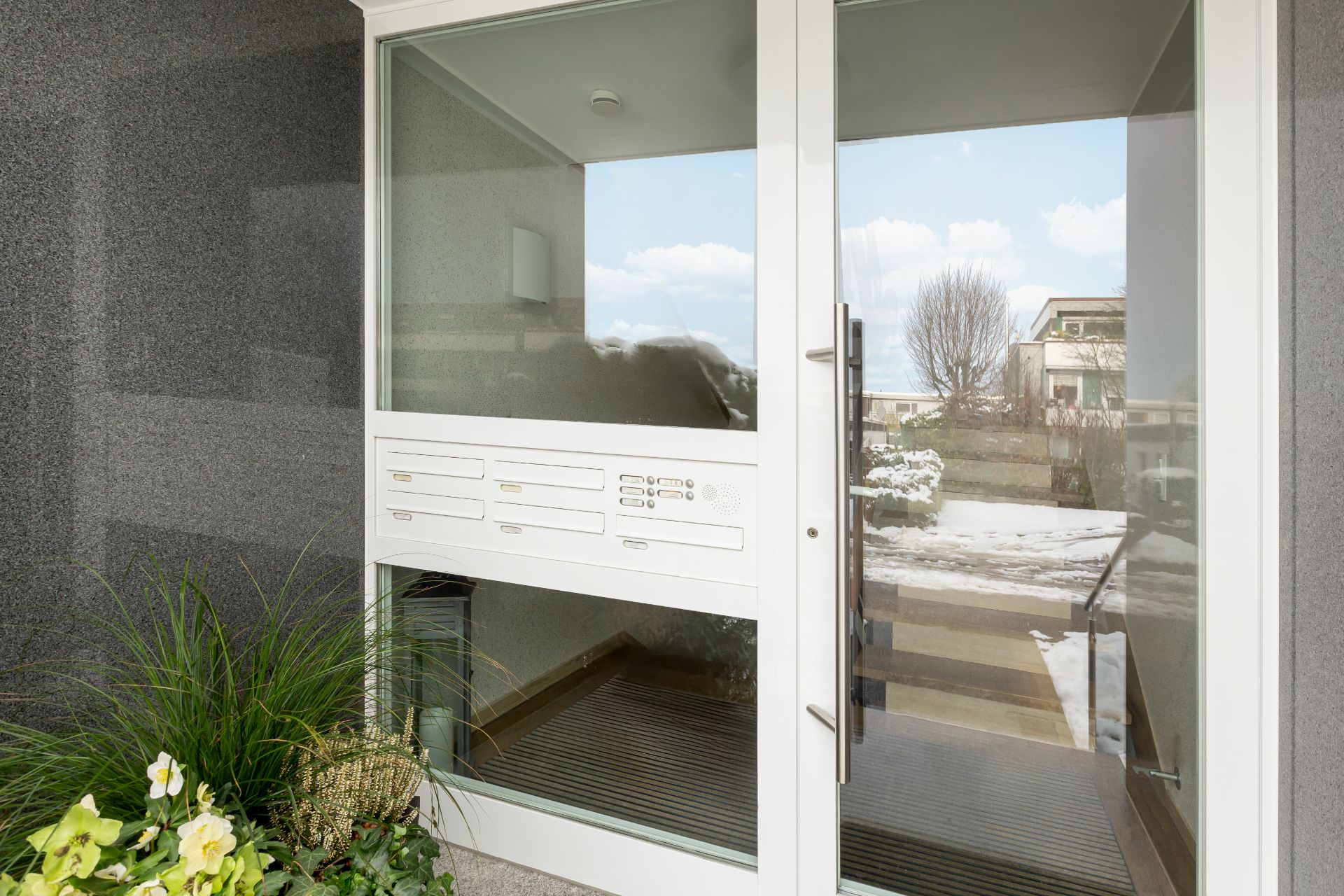 Hauseingang mit Glasfront, Briefkästen und Pflanzen neben der Tür.