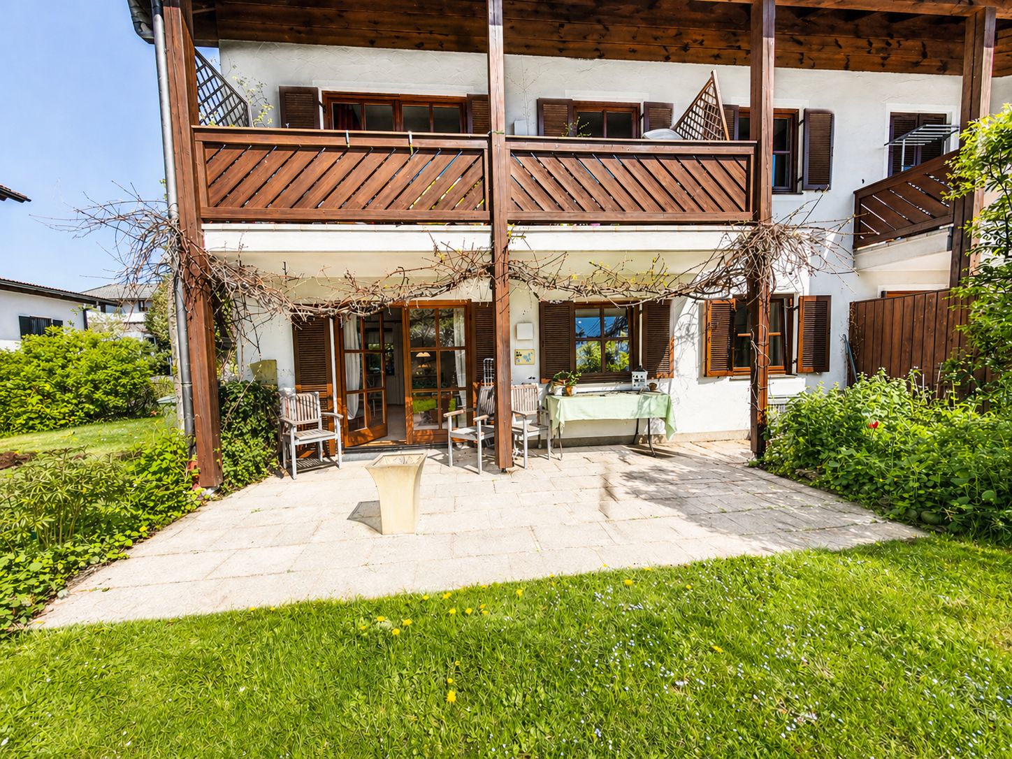Terrasse mit Sitzmöbeln vor einem weißen Wohnhaus mit Holzbalkon und Garten.
