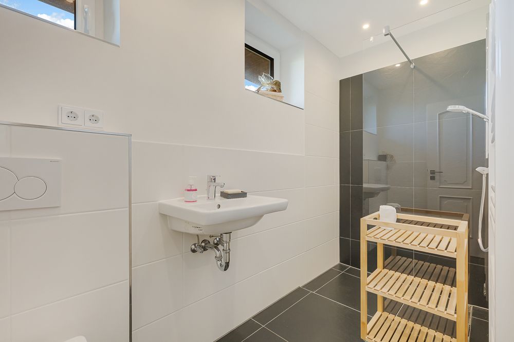Modernes Badezimmer mit Waschbecken, begehbarer Dusche und Holzregal auf dunklem Fliesenboden.